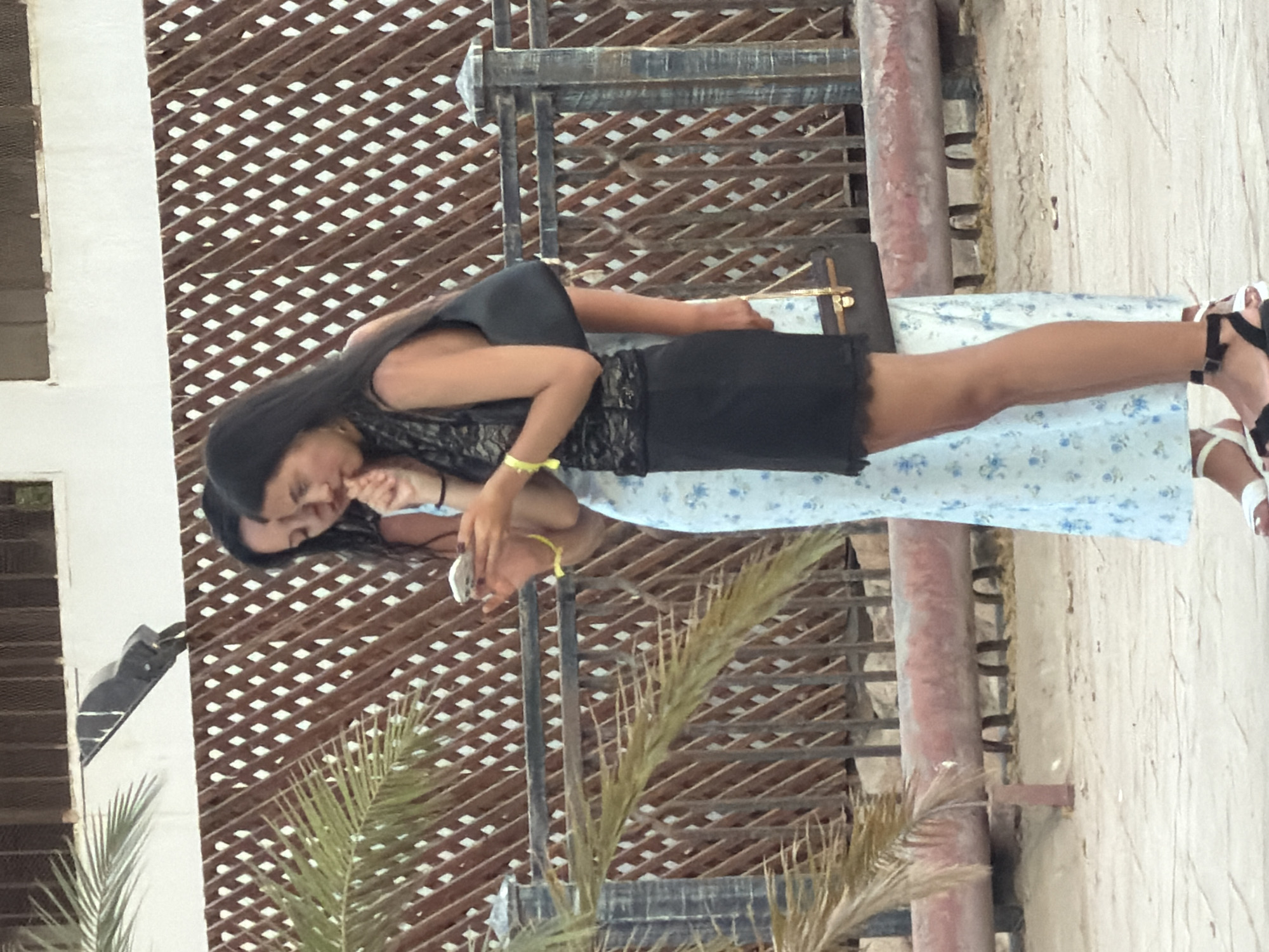 Two young women stand outdoors. The woman on the left wears a pale blue dress with a floral pattern. The woman on the right wears a black lace top and a black skirt. She holds a black handbag over her left shoulder and a smaller brown handbag in her right hand. Both women are looking down at a smartphone held by the woman on the left. They are standing in front of a brown wooden lattice fence and a metal railing. A palm tree branch is visible on the left side of the image. The ground appears to be sand or dirt. The lighting suggests it is daytime.