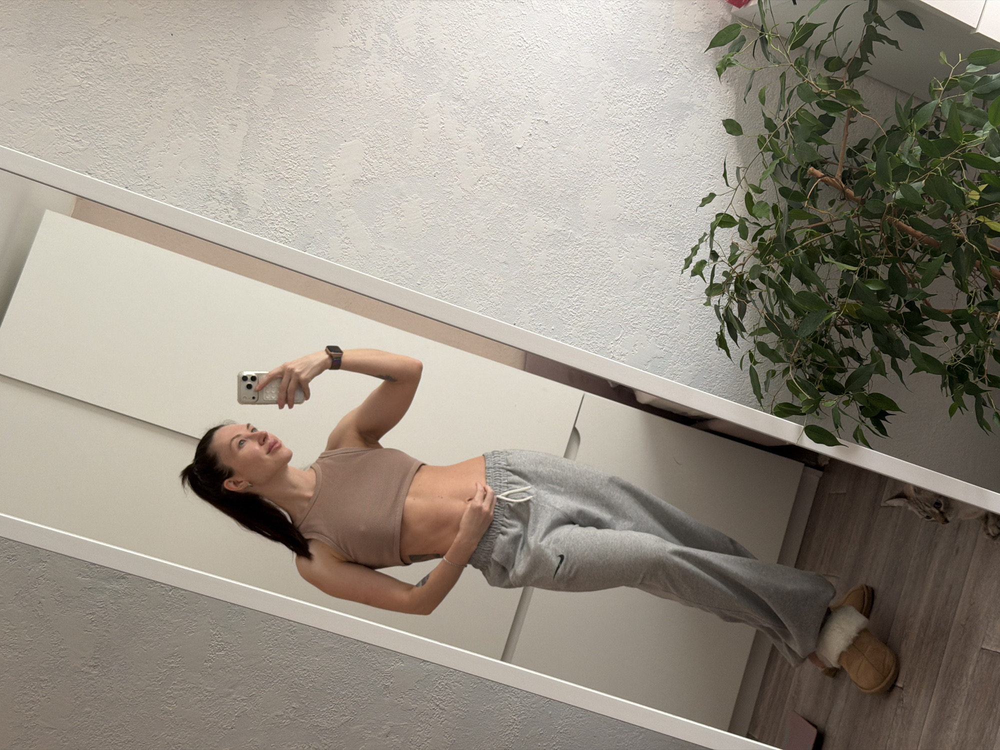 A woman stands in front of a mirror taking a selfie. She wears a beige tank top and gray sweatpants. She has her hair in a ponytail. She is wearing a smart watch on her left wrist. The reflection shows slippers on the floor. A plant sits in the corner. A cat is hiding beneath the plant. The wall behind the woman appears to be textured. The wall in the reflection appears to be a smooth surface. There is no indication of the location being Perm, Russia.