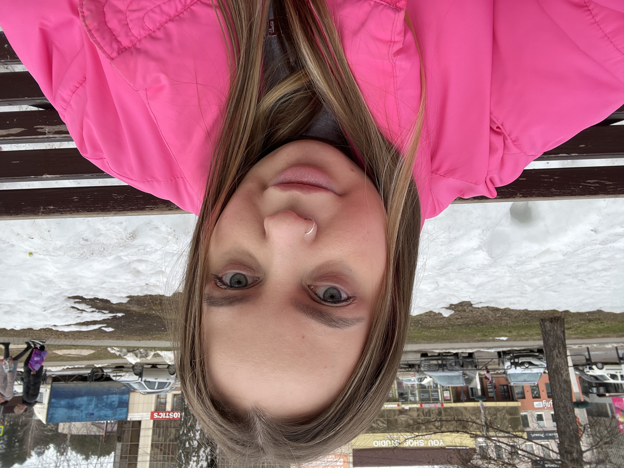 A female individual with light brown hair, direct eye contact with the camera, and a silver nose ring on her left nostril is positioned in the foreground. She wears a bright pink padded jacket over a grey top. She is seated on a dark brown slatted wooden bench, visible behind her right shoulder and arm. The immediate background consists of ground partially covered by melting snow and exposed dark earth.
Beyond this, a paved street runs horizontally across the midground. Parked vehicles on the far side of the street include a white van, a silver SUV (Renault Kaptur/Duster type), and a dark grey SUV. Buildings line the street, with one displaying "YOU SHOP STUDIO" in yellow lettering. Further to the right, a large blue box truck with a white cabin is visible, along with a partial red sign displaying "ROSTICS." Two pedestrians, one carrying a purple bag, are walking on a sidewalk parallel to the street. The scene depicts an urban streetscape in Chaikovsky, Russia, during a period of snowmelt.