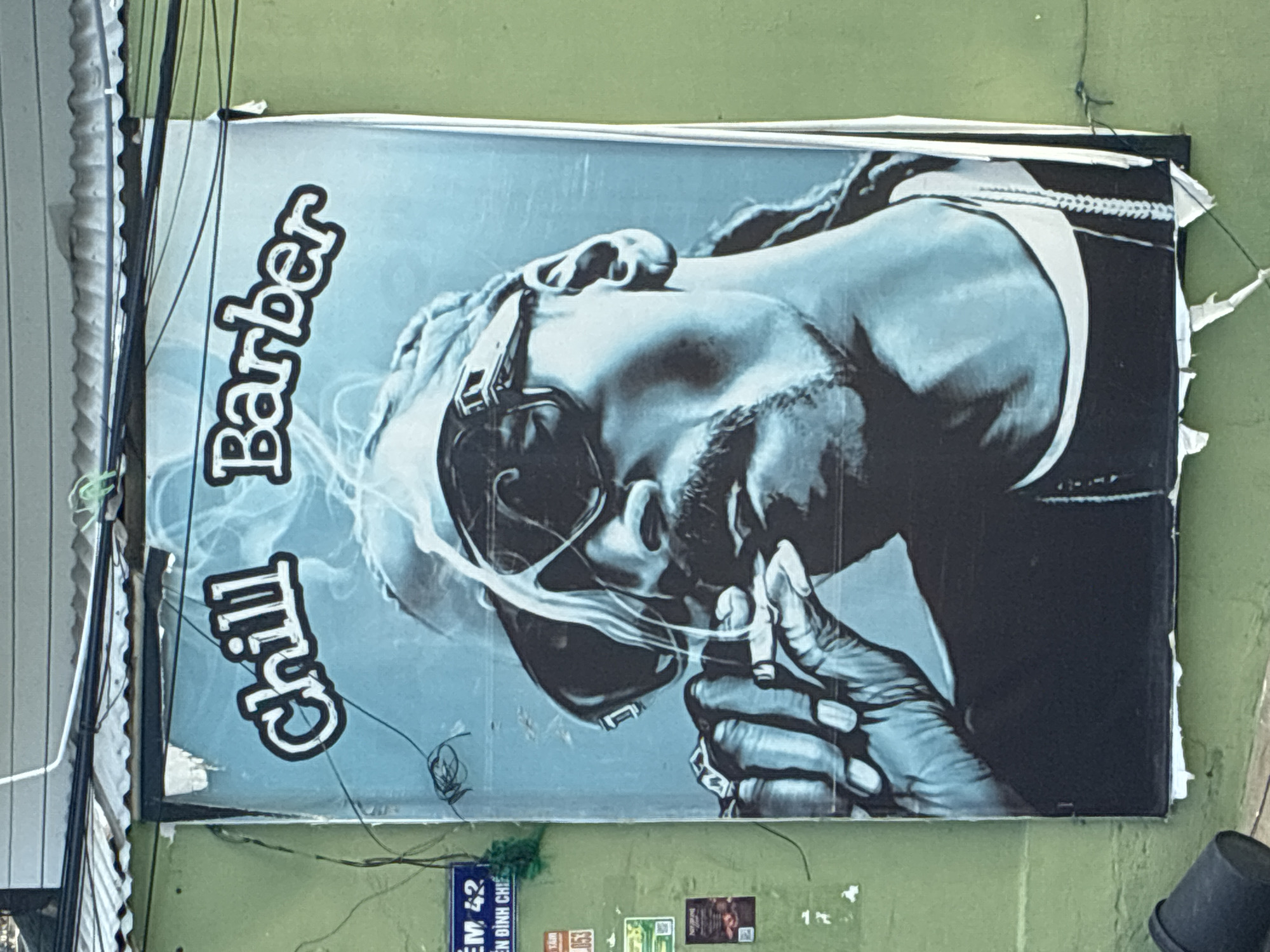 The image shows a large poster of rapper Snoop Dogg smoking a cigarette, with the words "Chill Barber" printed above him. The poster is affixed to a green wall in a city. The poster is partially torn and weathered. On the left side of the poster, there is a blue street sign with white text and a smaller orange sign. The street sign reads "EM 42 NGUYEN DINH CHI". A black bucket is visible at the bottom left of the image. The poster is hanging outdoors, with wires and a corrugated metal roof visible above it. The scene appears to be on a street in Bac Nha Trang Ward, Vietnam.