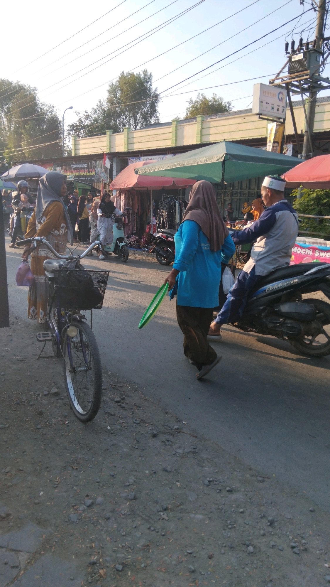 The image depicts a street scene in Tegalwangi, Indonesia, likely during the daytime with clear skies. A woman wearing a brown hijab and a turquoise shirt is walking away from the camera, holding a green ring. To her left, a bicycle with a basket is parked on the side of the road. Further in the background, a man wearing a blue and grey jacket and a white cap is riding a black motorcycle. Several market stalls with colorful awnings are visible, displaying clothing and other goods. Other individuals are also present, some on scooters and others browsing at the stalls. The ground is paved with asphalt and dirt, with some scattered debris. A utility pole with electrical equipment stands on the right side of the image.