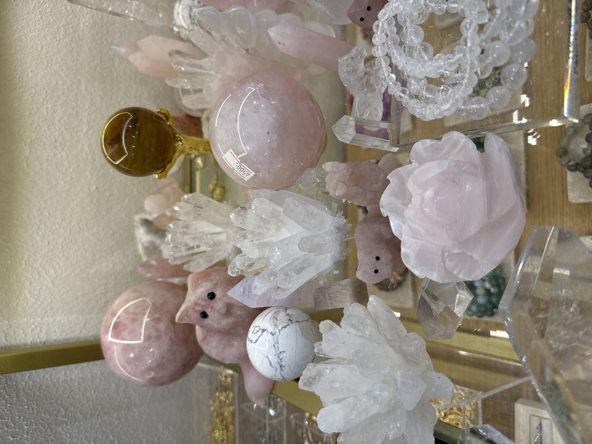 This media file features a close-up, eye-level shot of a display of polished and raw gemstones and crystals. The subjects are primarily objects, including spheres, clusters, and carved figures of various stones like rose quartz, tiger's eye, and clear quartz. There are also at least two carved stone figures that appear to be small, pink pigs with black bead eyes. A prominent rose quartz sphere has a white sticker with black text and numbers. A tiger's eye sphere is held by a golden talon-like fixture. The arrangement suggests a retail or display setting, possibly a shop specializing in crystals or New Age items. The lighting is bright and appears to be artificial, creating reflections on the smooth surfaces of the polished stones. There are no visible people, and the activity is static, focusing on the presentation of the items. The background is out of focus, showing textured surfaces and hints of wooden shelving. The context provided indicates the location is Saint Petersburg, Russia. The visible text on the rose quartz sphere reads "4500.".