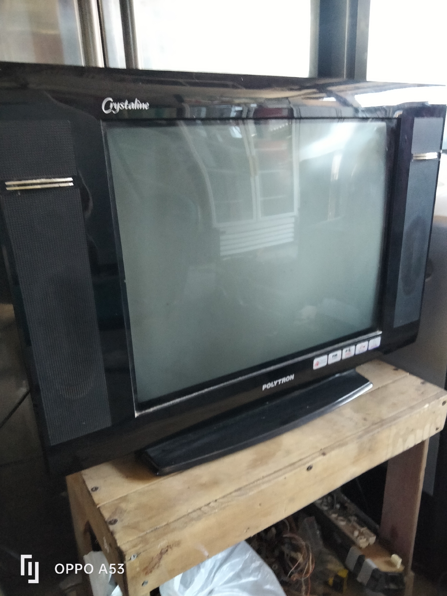 A black CRT television is placed on a wooden stand. The television has a screen that reflects light and shows some interior details. The brand "Crystalline" is visible on the top left of the television, and "POLYTRON" is printed on the bottom front. There are several buttons on the lower front panel of the television. The television is situated indoors, with a blurred background suggesting an indoor setting. The stand is made of unfinished wood planks, and beneath it, some discarded items and wires are visible. The lower right corner of the image contains a watermark that reads "OPPO A53". The image appears to have been taken in Denpasar, Indonesia.