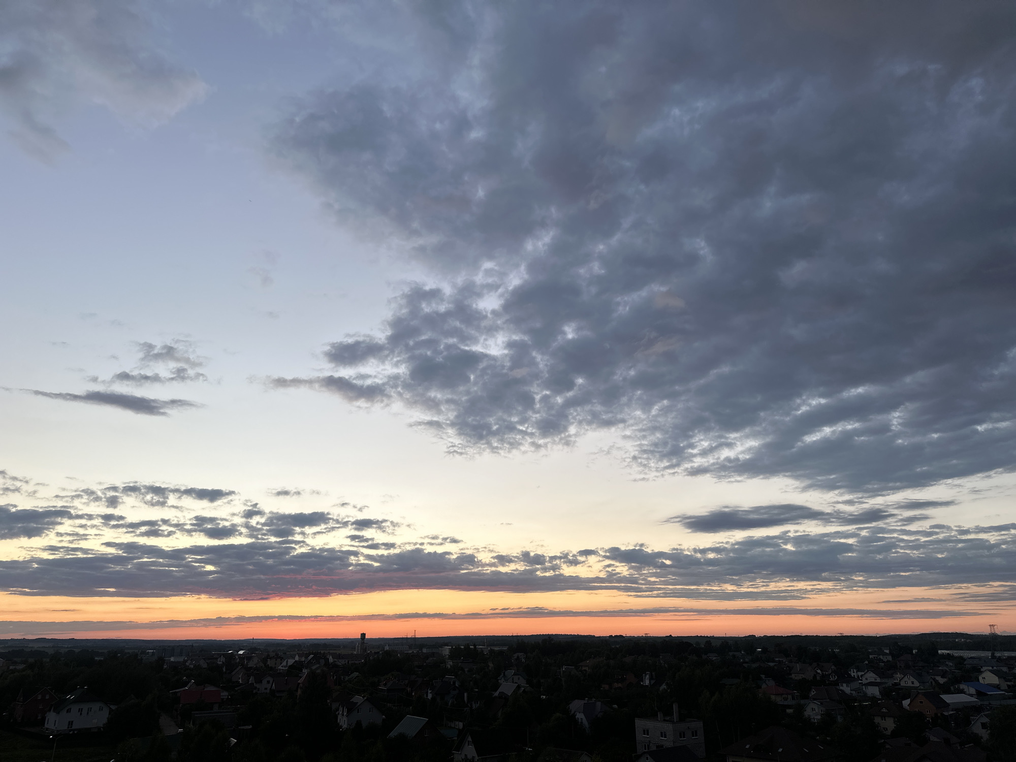 The image depicts a panoramic view of a suburban area at dusk, likely from an elevated vantage point. The sky is dominated by a gradient of colors, transitioning from a pale blue at the top to vibrant orange and pink hues near the horizon, indicating the setting sun. A layer of dense, dark gray clouds partially obscures the sky, creating a dramatic contrast with the warmer tones of the sunset. Below the sky, a sprawling landscape of residential buildings and dense foliage stretches across the frame. The rooftops of houses are visible, clustered together, interspersed with dark green trees. The overall impression is one of a quiet evening settling over a residential neighborhood. The location is identified as the Shchomyslitsky Sel' soviety, Belarus.
