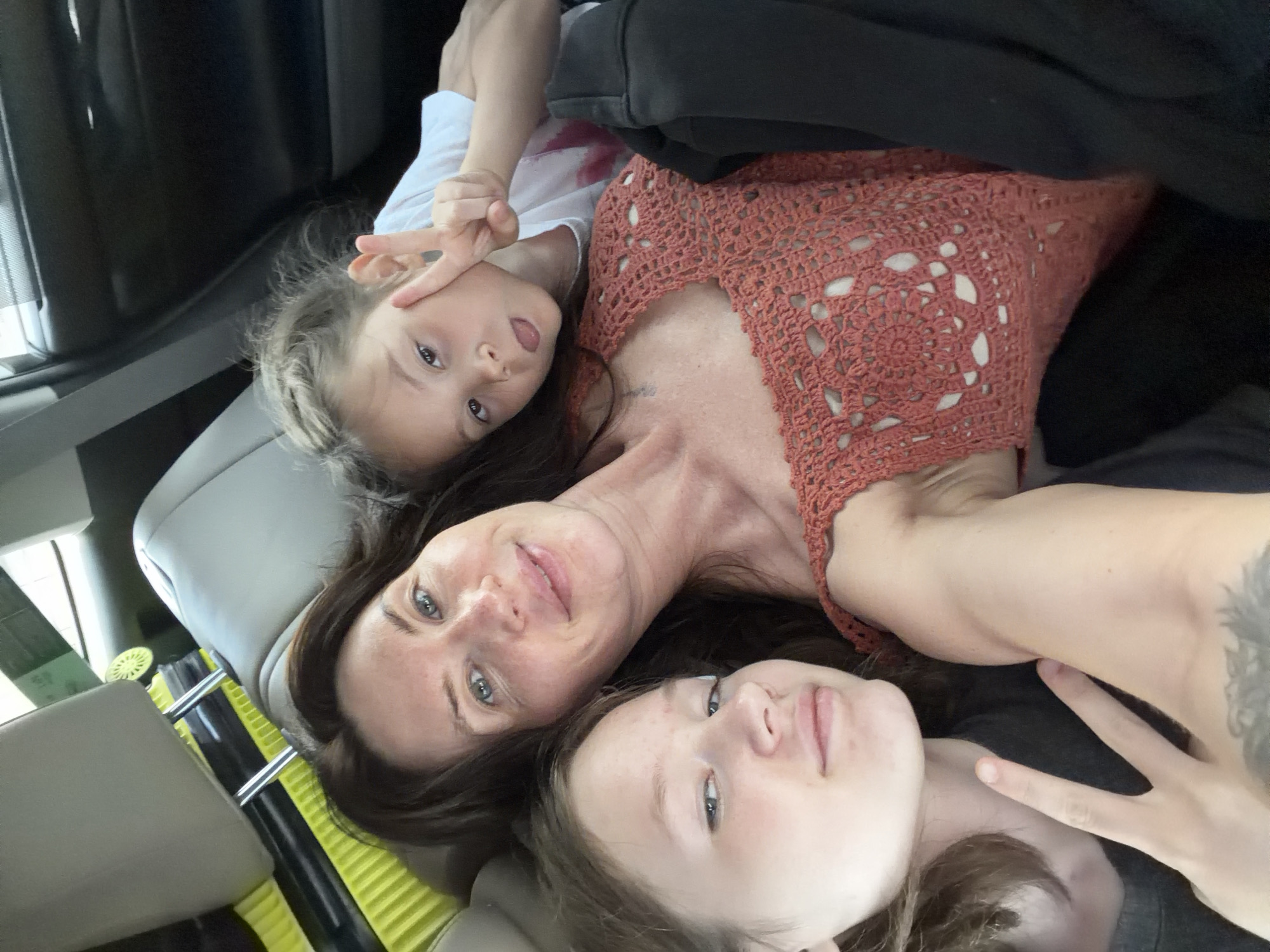 An overhead, close-up shot shows a woman and two children in the back seat of a vehicle. The woman has long, dark brown hair and is wearing a coral-colored, crocheted top. She is looking at the camera with a slight smile. To her left is a child with brown hair, who is also looking at the camera and holding up a peace sign. To the woman's right is another child with brown hair, who is sticking out their tongue and holding up a peace sign. The children appear to be young. The background shows the interior of a car, including car seats and luggage. The lighting suggests it is daytime. The image does not contain any information about the location in the city of Ильинское, Russia.