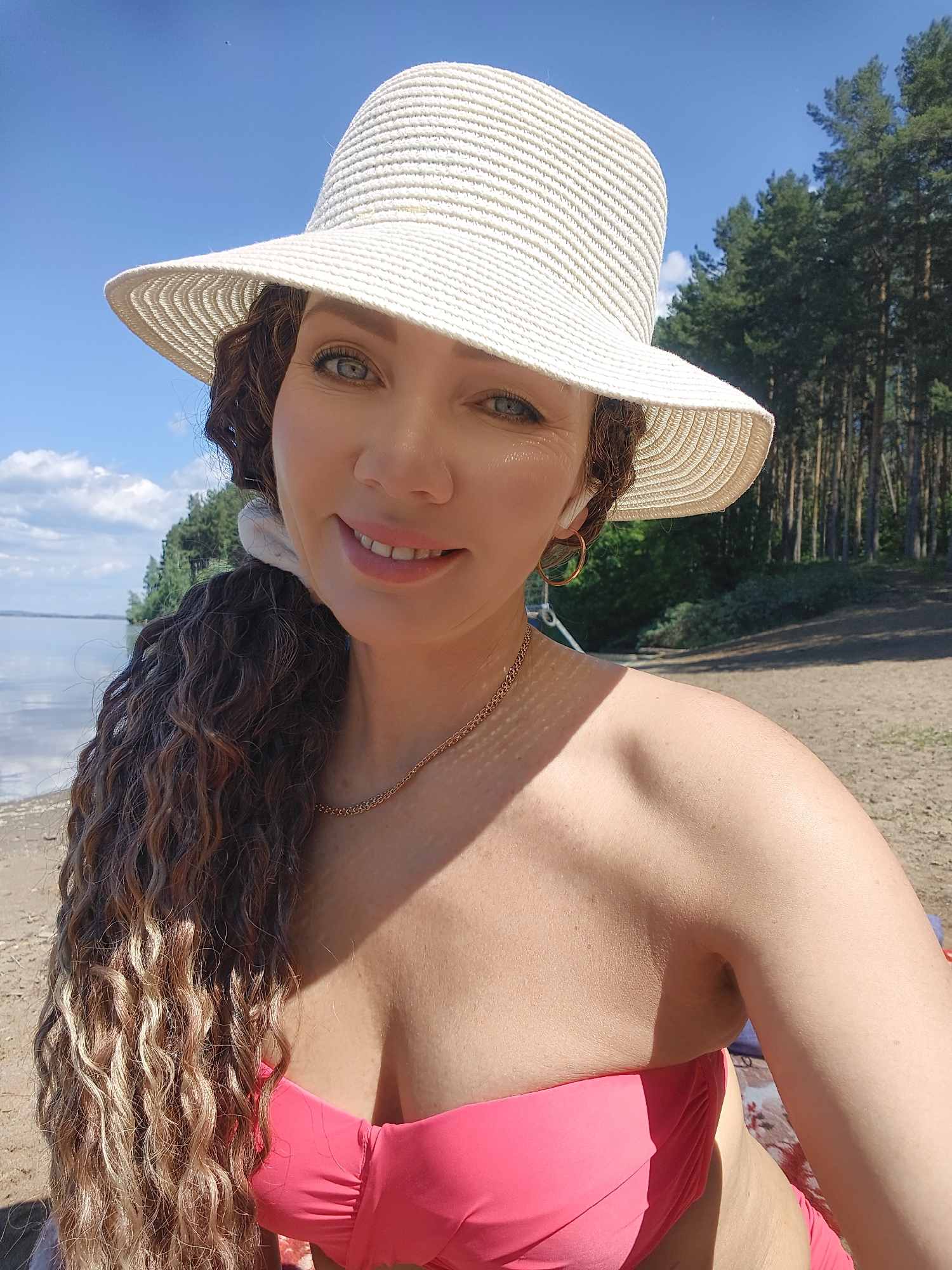 A woman is seen from the chest up, wearing a white straw hat and a pink bikini top. She has long, curly brown hair and is wearing a gold chain necklace and a hoop earring. She is on a beach with a body of water and trees in the background, under a clear blue sky with some clouds. The woman is smiling and looking towards the camera.