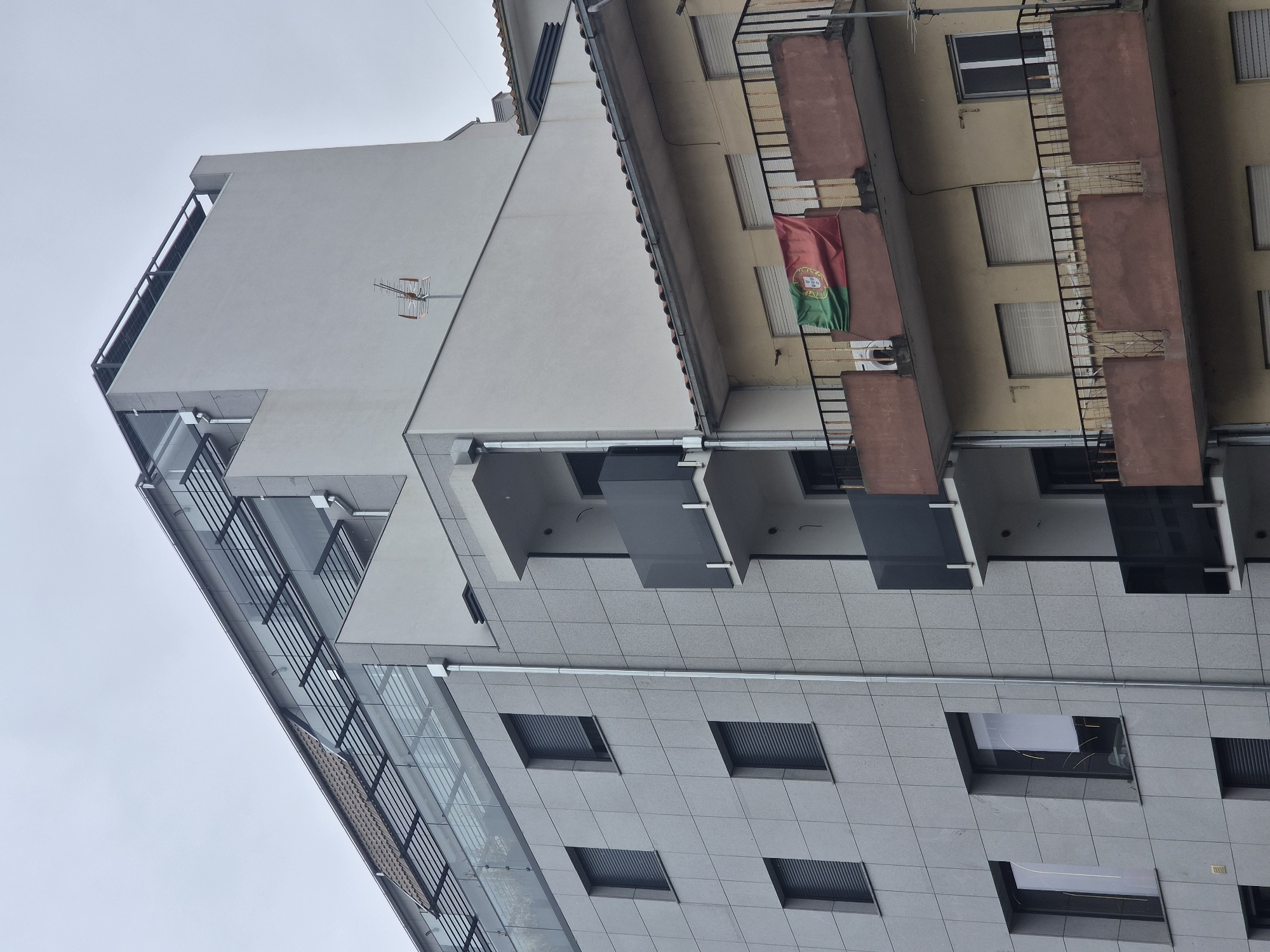 The image shows a close-up, slightly low-angle view of the exterior of two adjacent multi-story buildings in Bragança, Portugal, under an overcast sky. The building on the left is modern, clad in large, light grey rectangular tiles, and features several windows with closed roller shutters. Above the tiled section, there's a glass-enclosed upper level with a sloped, tiled roof segment. The building on the right is older, with a yellowish facade and multiple balconies. A Portuguese flag is prominently displayed, draped over the railing of a balcony on the upper floor of the older building. The balconies of the older building have dark metal railings and reddish-brown facing panels. A satellite dish or antenna is visible on the roof of the modern building. The overall impression is urban residential architecture.