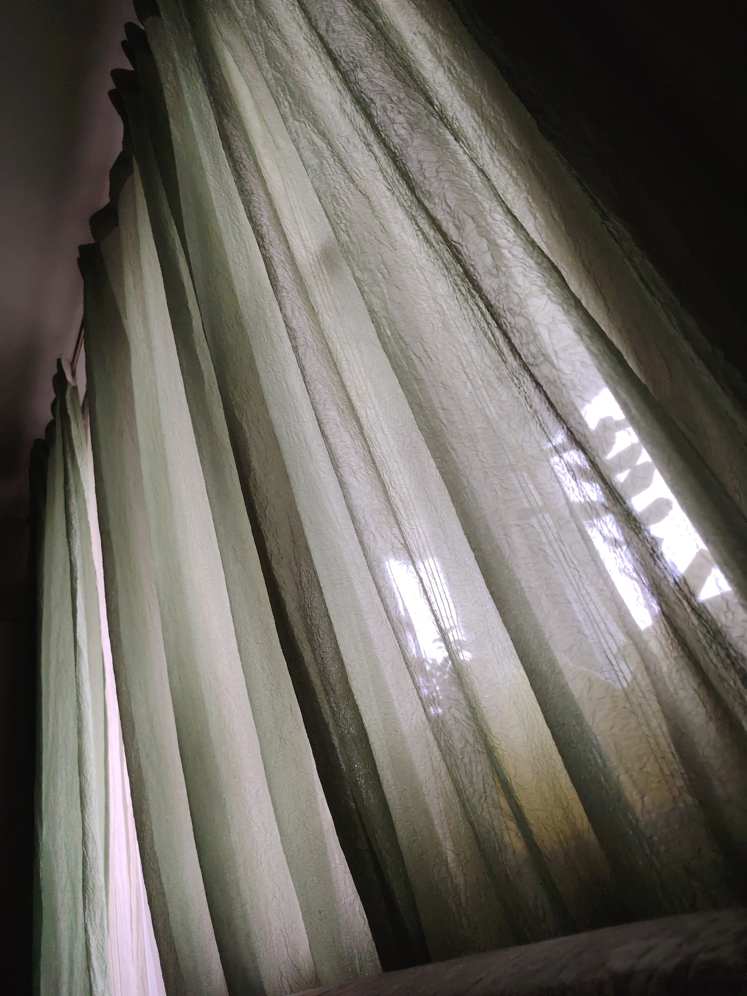 The image shows a close-up, upward-angled view of sheer, light-green curtains hanging in a room. Light filters through the curtains, suggesting a window behind them. The texture of the curtains is visible, with a wrinkled or slightly patterned appearance. The room's ceiling and wall are visible at the upper left, suggesting a dark interior. It is not possible to verify the location in Chaikovsky, Russia based on the provided image.