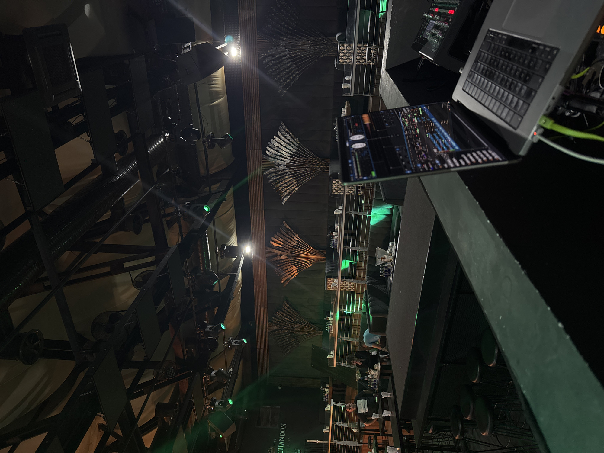 This image captures the interior of a dimly lit club or event venue in Tsarevo, Bulgaria, bustling with activity at night. The scene is dominated by a DJ booth on the right, featuring a silver Apple MacBook Pro laptop displaying colorful audio waveforms or visualizer software. Adjacent to the laptop is an audio mixer or controller with multiple knobs and faders, hinting at a live music or entertainment event.
The ceiling features an industrial design with exposed dark metal beams, ventilation ducts, and various stage lights, some emitting green glows. Several decorative elements resembling fanned-out, dried natural materials (possibly leaves or reeds) are suspended from the ceiling, adding an artistic touch to the otherwise utilitarian structure. Large speakers and sound equipment are integrated into the overhead framework.
In the background, patrons are visible seated at tables and on couches, appearing relaxed and socializing. One person in a dark t-shirt is standing near a table, while others are partially obscured. On the tables, drinks, glasses, and white napkins can be seen. A brand name, "CHANDON," is faintly visible on a sign or bottle in the background, suggesting champagne or sparkling wine is served. The overall atmosphere is dark and vibrant, characteristic of a nightlife setting, with focused lighting creating dramatic effects and highlighting specific areas.