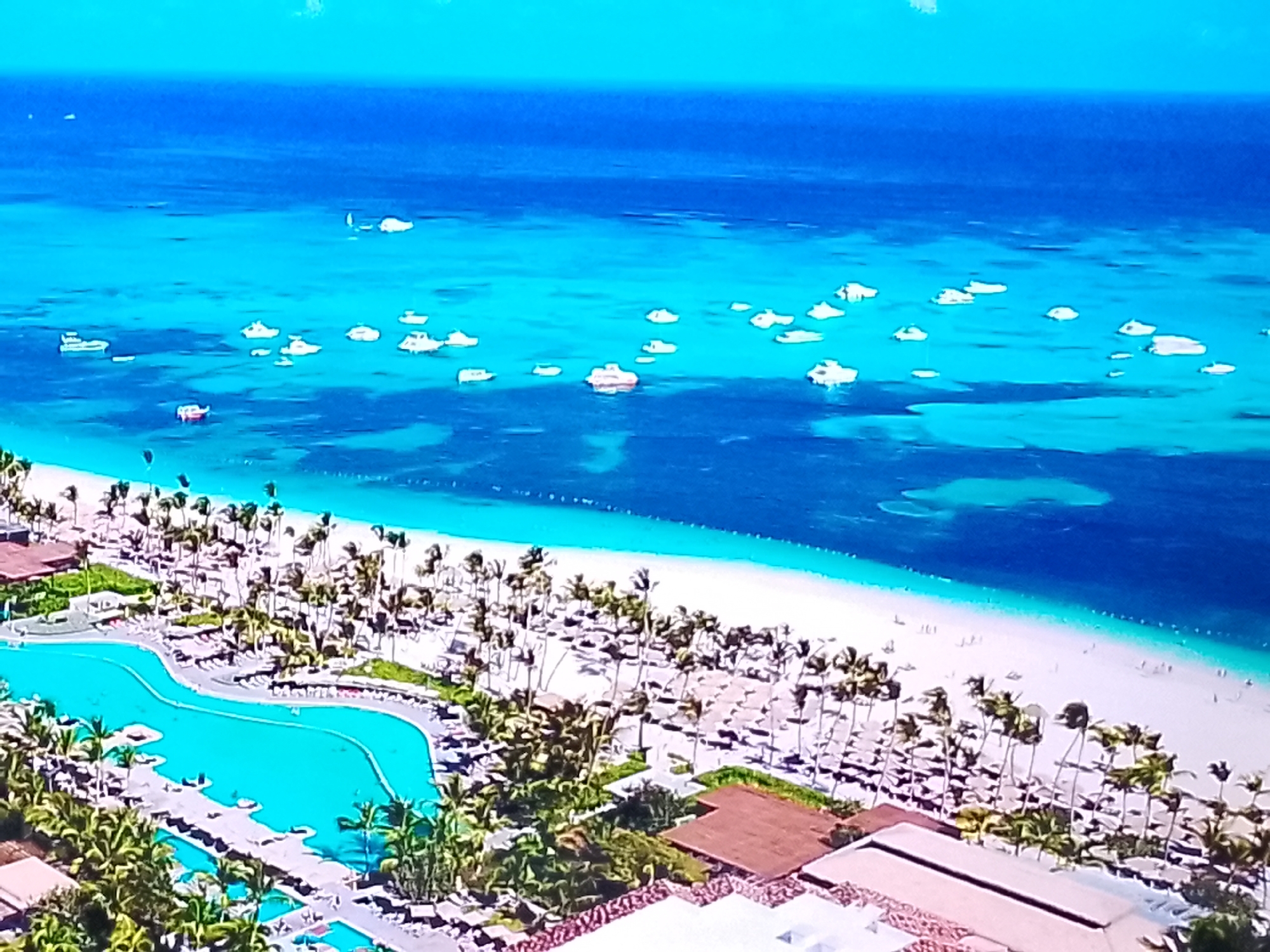 This is an aerial, wide-angle shot of a tropical beach resort. The foreground features a large, irregularly shaped swimming pool, surrounded by lush green palm trees and resort buildings with reddish-brown tile roofs. Further back, a wide expanse of white sand beach is dotted with numerous palm trees and scattered beach chairs. In the distance, the turquoise ocean meets a deep blue sea. Several small white boats are scattered across the water, some appearing to be moored. The sky is a clear, bright blue. The scene depicts a serene and idyllic vacation setting. There is no visible indication that this location is in the city of Chișinău, Moldova.