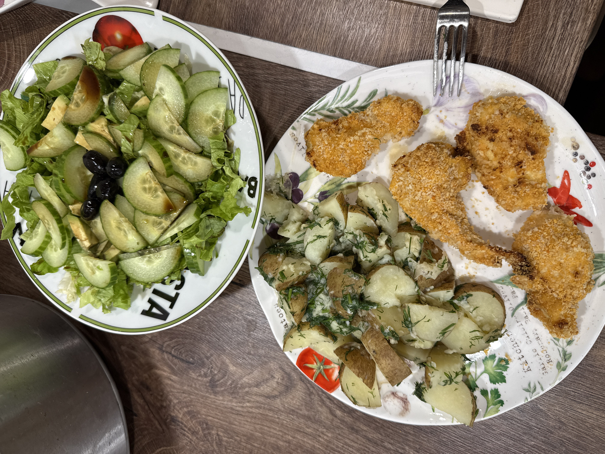On a wooden table, two plates hold food. The plate on the left contains a salad of lettuce, sliced cucumber, avocado, and black olives. The plate on the right holds boiled potatoes with dill and what appears to be breaded chicken or fish. A fork rests on the plate with the chicken and potatoes. No people are visible. The location is unknown, but is listed as Sofia, Bulgaria.