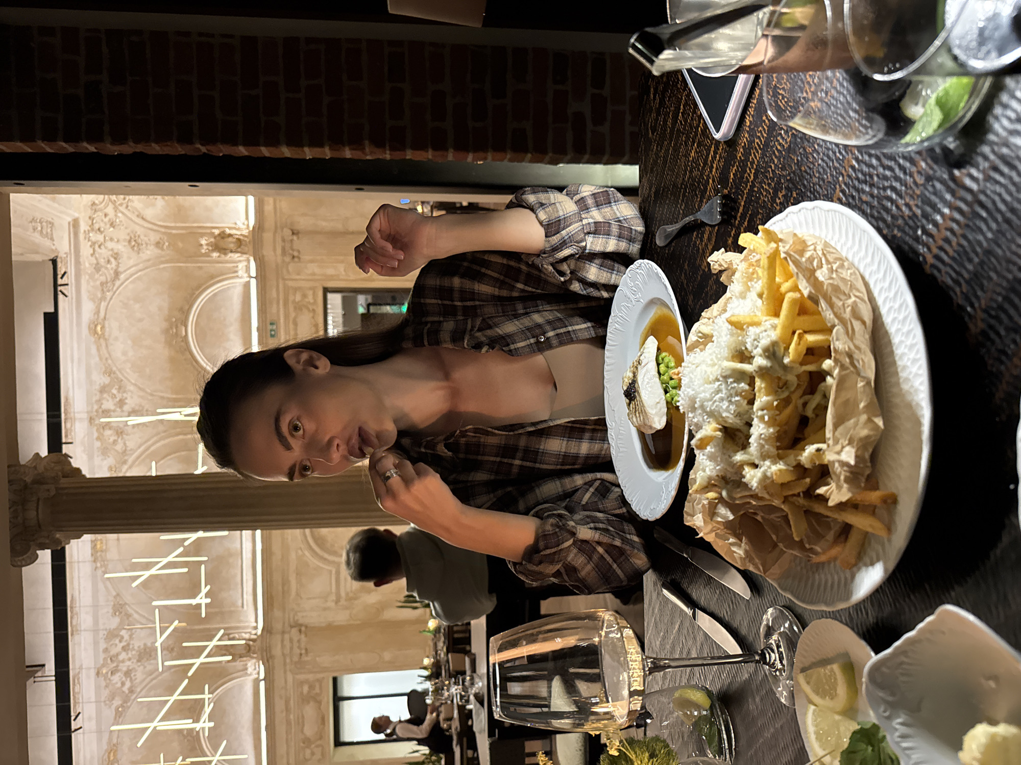 The image captures a seated woman at a dark, textured table in an upscale restaurant, likely indoors. The background showcases opulent architecture with a prominent column featuring an ornate capital, gilded scrollwork on walls, and arched alcoves, complemented by modern, linear light fixtures suspended from the ceiling. A section of a dark reddish-brown brick wall is visible on the far right.
The woman, estimated to be in her 20s-30s, has dark hair and wears a brown and beige plaid long-sleeve shirt over a light beige undergarment. A ring is visible on her right ring finger. She is looking slightly to her right, with her lips parted and her right hand raised to her mouth, holding a small object, suggesting she is about to consume or taste food.
On the table in front of her are several dishes and beverages. A significant portion of french fries, served on a white plate and wrapped in brown paper, is heavily topped with a white creamy sauce and what appears to be grated cheese or white flakes. To its left, another white plate holds a main course: a piece of white fish adorned with black caviar, resting on green peas, all immersed in a dark brown sauce. A clear wine glass, partially filled with white wine, is positioned to her left. Additionally, a small white dish contains two lemon wedges and green herbs, and a small white bowl holds a yellow substance, possibly butter. Silverware, including two knives and a fork, is arranged on the table. A dark-colored smartphone with a light-colored case, a copper-colored container, and another clear glass containing a drink with a lime wedge are visible on the right side of the table.
In the blurred background, another diner wearing a grey hooded sweatshirt is seated, facing away from the camera. Further in the distance, a restaurant staff member in a dark uniform with a white top is visible. The scene is illuminated by dim, ambient lighting, creating a refined atmosphere.