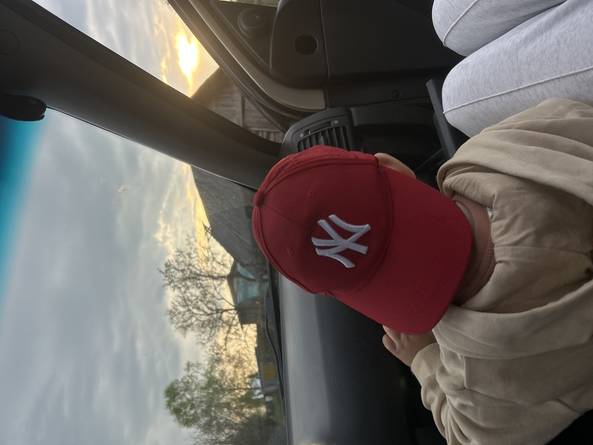 A view from inside a car in Малая Борщевка, Russia, at dusk. A child wearing a red New York Yankees baseball cap and a beige hoodie is seated. Another person is seated, wearing grey sweatpants. Through the windshield and the open car door, there is a view of rural scenery, including trees, a house, and an overcast sky with a hint of sunset.