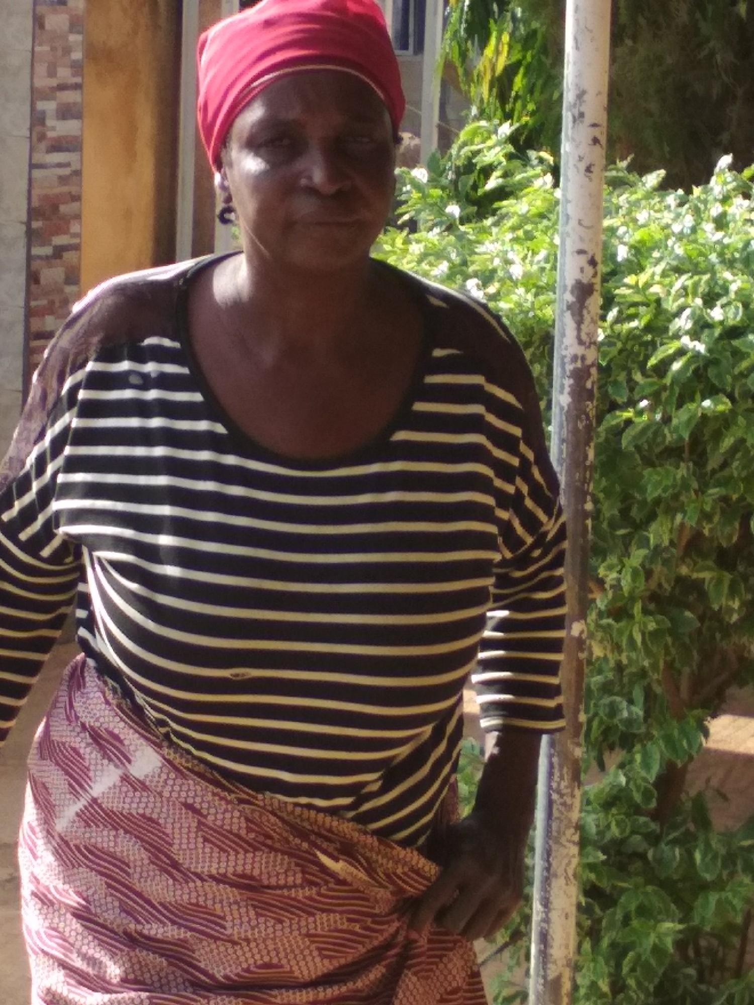 A medium shot captures a dark-skinned woman standing outdoors in Barakin Sabawa, Nigeria. She appears to be of middle age or older and looks directly at the viewer with a neutral, somewhat serious expression.
The woman is wearing a vibrant red headscarf that covers her hair. Her top is a long-sleeved shirt with alternating black and light-colored (possibly white or cream) horizontal stripes. Around her waist, she has wrapped a patterned piece of fabric, predominantly reddish-brown with intricate lighter geometric designs. Her right hand is partially visible near her hip, lightly holding the wrapper.
The setting appears to be outdoors during daytime, with bright natural light causing some glare on her forehead and cheek. To the woman's right, a weathered, light-colored pole, possibly metal, stands vertically. Behind this pole and extending to the right is a dense patch of green foliage, likely bushes or small trees, some bearing small white flowers. To her left, behind her, part of a building with a textured, brick-like wall is visible.
No specific activity is taking place; the woman is standing still, posing for the photograph. There is no visible text in the image.