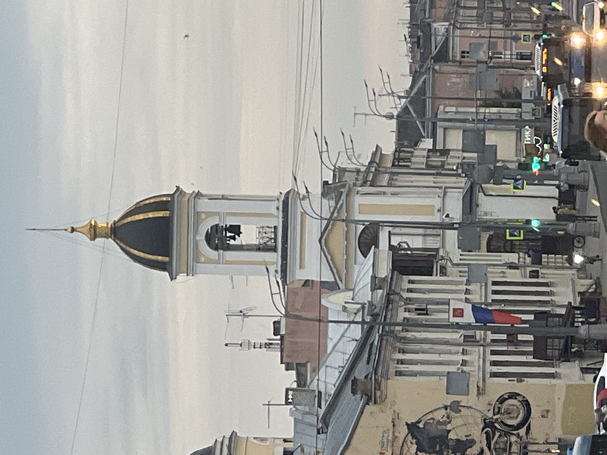 The image shows a city street scene, likely in Tver, Russia, given the presence of a Russian flag. A prominent bell tower with a golden dome stands tall, a religious structure that is characteristic of many Russian cities. The tower has multiple openings where bells are visible. Below the tower, a street with traffic and pedestrians unfolds. Cars, including what appear to be buses, are on the road, some with illuminated headlights. Streetlights and traffic signals are also visible. The buildings lining the street are multi-story, exhibiting various architectural styles. One building features a mural depicting a motorcycle and a world map. The sky is overcast, suggesting a cloudy day. The overall impression is that of a typical urban environment in Russia.