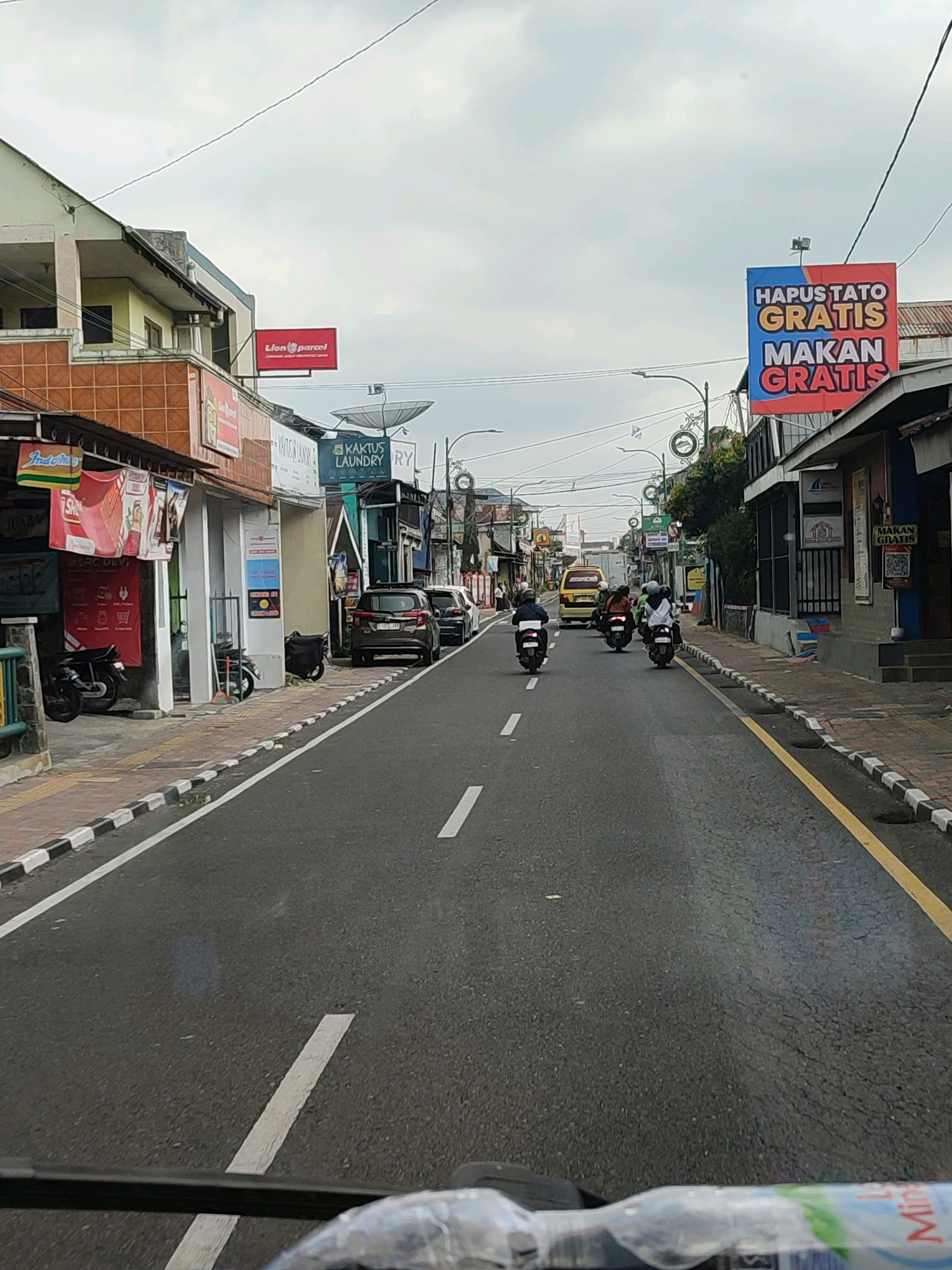 The image shows a street scene in Jaraksari, Indonesia. A road runs through the middle of the frame, with buildings on either side. A number of motorbikes and cars are visible on the road, as well as a yellow van. A banner on the right side of the road reads "HAPUS TATO GRATIS MAKAN GRATIS". The sky is overcast.