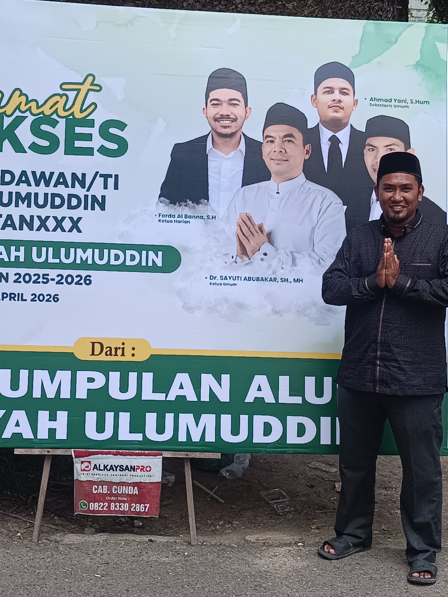 The image depicts a man standing in front of a banner. The banner features the text "SELAMAT SUKSES" (Congratulations on Success), followed by details about a group called "ALUMUDDIN," including their inauguration, the years 2025-2026, and the month of April 2026. The banner also shows the photos of a man dressed in a white outfit, likely the leader or a figure of authority for that organization. There are additional men in suits and ties behind him.
Beneath this information, the banner states, "Dari: KUMPULAN ALUMNIYAH ULUMUDDIN" (From: Group of ULUMUDDIN Alumni). Attached to the bottom of the banner is a sign for "ALKAYSANPRO," a paint services and event production company. The sign specifies "CAB. CUNDA" (Cunda Branch) and provides an order number: 0822 8330 2867.
The man standing in front of the banner is wearing a dark, patterned shirt, dark trousers, and sandals. He is smiling and his hands are pressed together.
The location is Lhokseumawe, Indonesia.