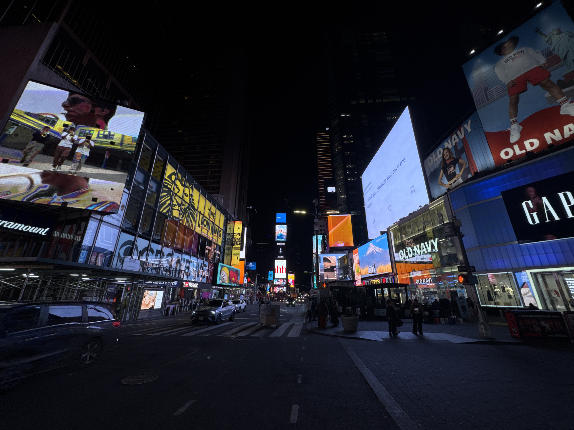 The image shows a street scene in Times Square, New York City, at night. Illuminated billboards line the streets, displaying advertisements for various brands, including Paramount, Old Navy, Gap, and Tissot. Pedestrians are walking on the sidewalks and crossing the street. Cars are traveling down the road. The overall impression is one of a busy, brightly lit urban environment at night.