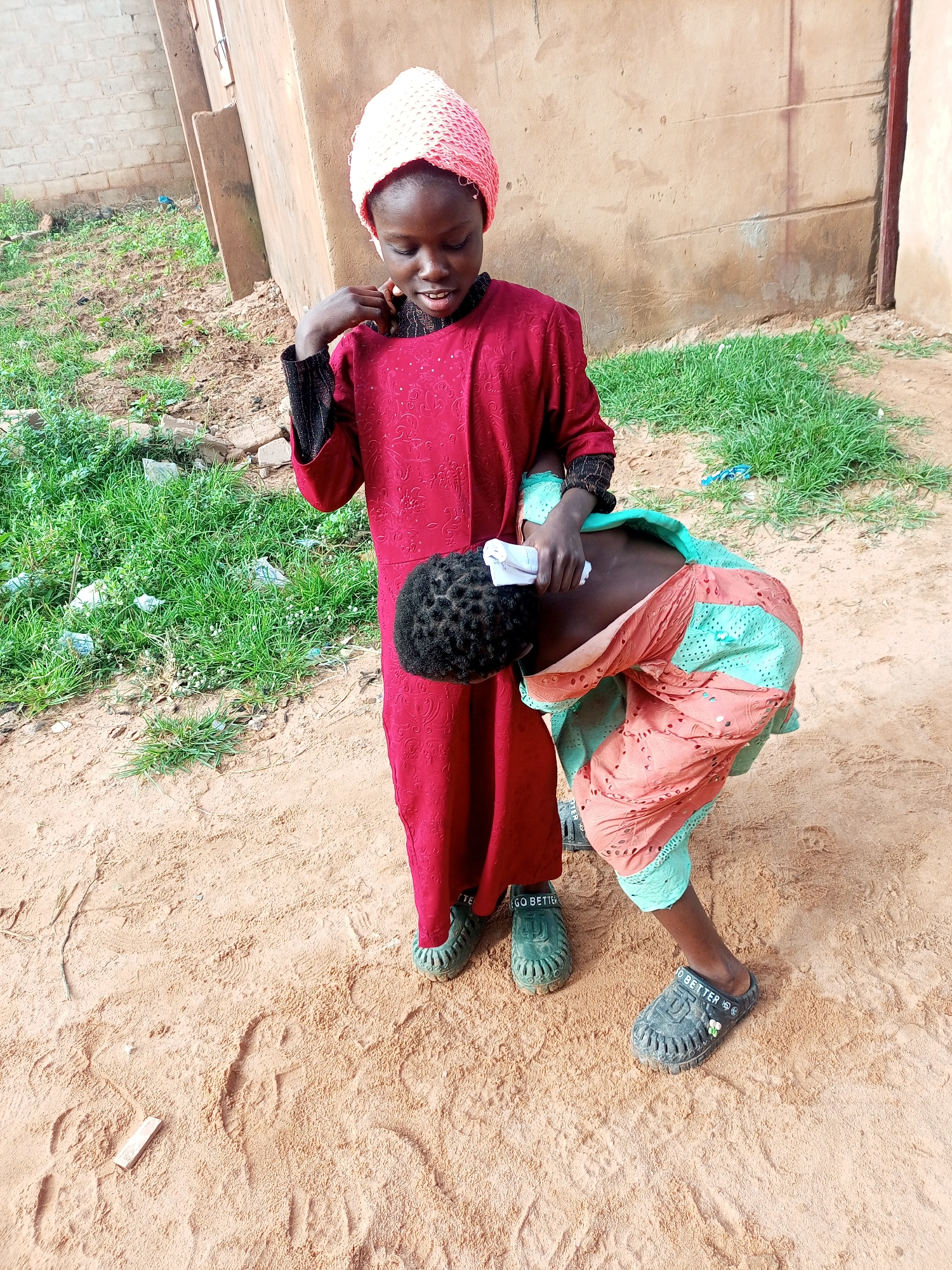 Two young girls are in an outdoor environment. The girl on the left is wearing a red dress and a pink headscarf. She is looking down and touching her neck. The girl on the right is bent over, with her back to the camera, wearing a light orange and turquoise patterned outfit. She is holding a white cloth to the back of her head, which is covered in dark curly hair. Both girls are wearing sandals. The ground is made of sand, and there is patchy green grass and some debris in the background. A tan-colored building is visible behind them. The scene appears to be a residential area in Tempure, Nigeria.