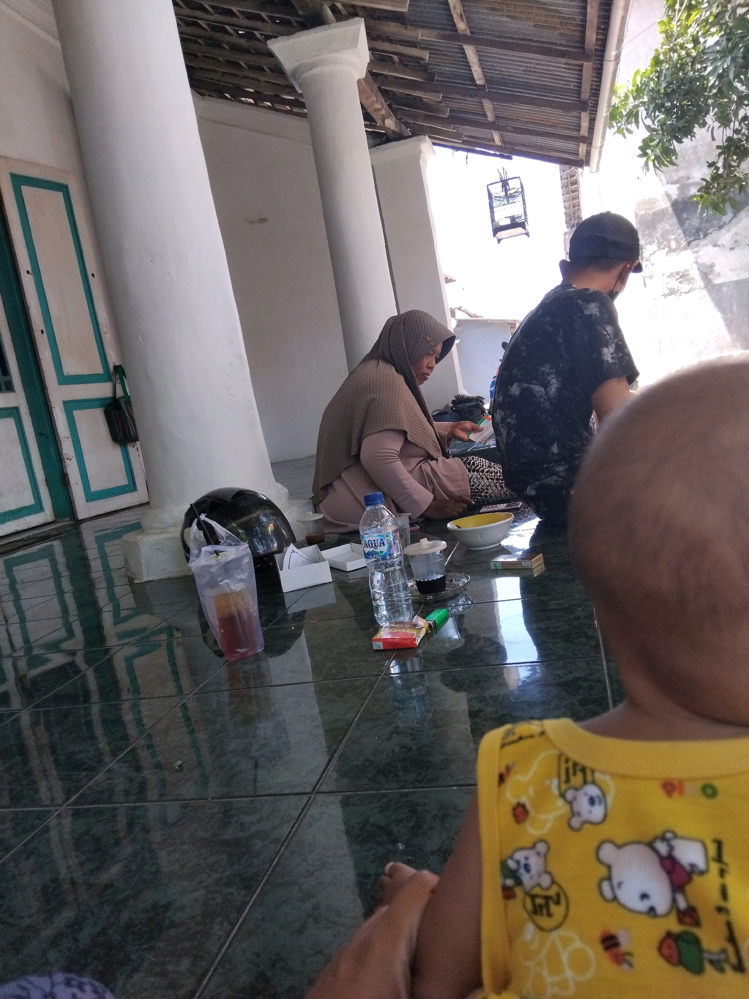 The image depicts an outdoor scene in Kepuh Kiriman, Indonesia, with a tiled floor reflecting the overhead structure. In the mid-ground, a woman wearing a brown hijab and a light brown outfit is seated, appearing to be engaged in an activity on a white box. To her right, a person in a dark t-shirt and a cap is also seated, facing away from the camera. A black helmet rests on the tiled floor near the woman. Several objects are scattered on the floor, including a clear plastic cup with a dark liquid and ice, a bottle of Aqua water, a white container, a pack of cigarettes, and a lighter. In the background, a birdcage hangs from the corrugated metal roof structure. The overall environment suggests a casual, possibly residential, outdoor setting.