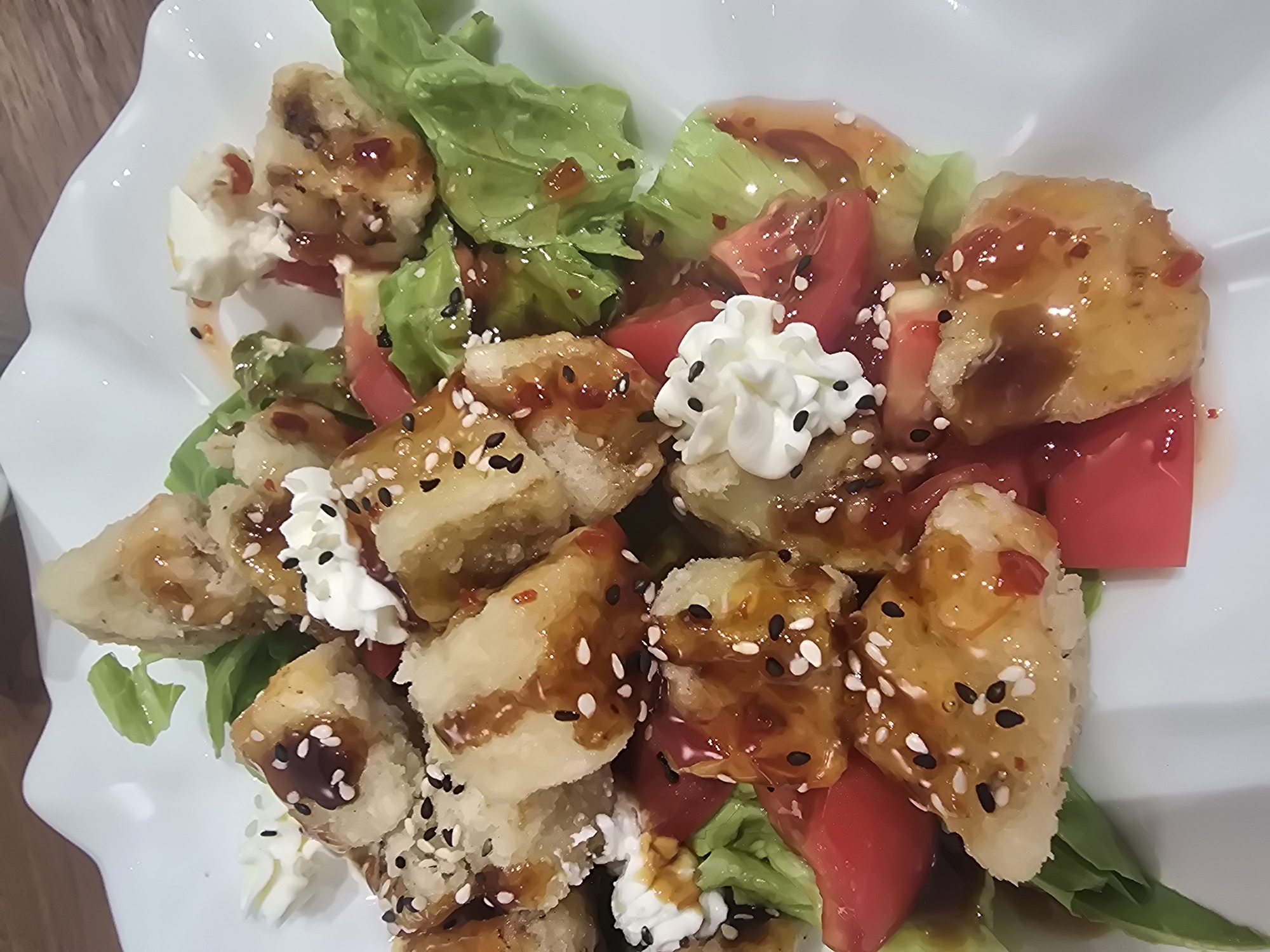This is a close-up, high-angle shot of a dish of fried food served on a white, fluted plate. The dish appears to be a salad with pieces of fried food, lettuce, and tomatoes. The fried pieces are golden brown and coated in a dark, glossy sauce, sprinkled with white and black sesame seeds. There are dollops of white cream or cheese scattered throughout the dish, some topped with black sesame seeds. The lighting is diffused, and the background is out of focus, showing a hint of a wooden surface. There is no information in the image to suggest a location in Ulan-Ude, Russia.