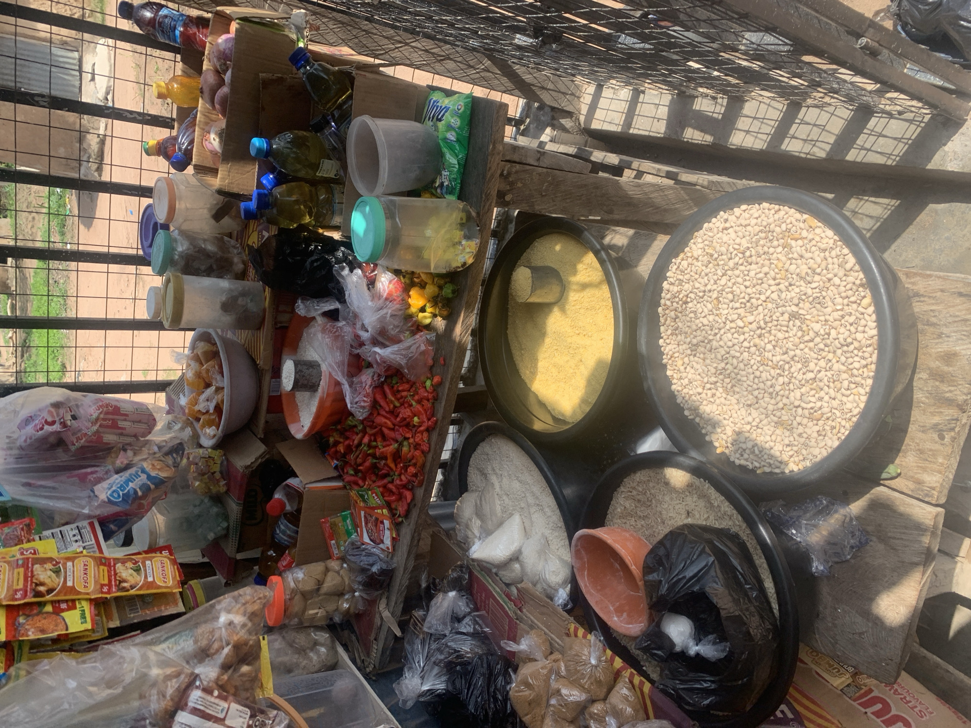 This is a medium shot, overhead, color photograph of an outdoor market stall in Jalingo, Nigeria. The stall appears to be a temporary setup with wooden planks and a metal grate overhead, casting a shadow on the ground. The focus is on a variety of goods for sale.
The subjects are primarily objects. There are several containers filled with various food items. Two large black bowls are prominent, one filled with what appears to be white beans and another with a yellowish grain, possibly cornmeal or semolina. Smaller containers hold red chilies, what looks like dried fruit or candy, and other unpackaged goods. Various bottles of liquids, likely cooking oils or beverages, are also visible, stacked on wooden shelves. Several packaged food items are present, including sachets of spices labeled "SANKOFA CHICKEN FLAVOUR" and a package with the text "JUMRO". Other items include plastic bags filled with various dry goods and what appear to be sausages or meat products.
The scene depicts a market stall stocked with staple food items and spices, typical of a local market in Nigeria. The lighting suggests it is daytime, with bright sunlight casting strong shadows, indicating it could be mid-morning or early afternoon. The presence of a metal grate overhead suggests an open-air market setting. There are no people visibly in the frame, and the overall impression is one of abundance and variety of goods for sale. The visible text "SANKOFA", "CHICKEN", "JUMRO", and "STRAWBERRY FLAVOUR" provides specific product information.