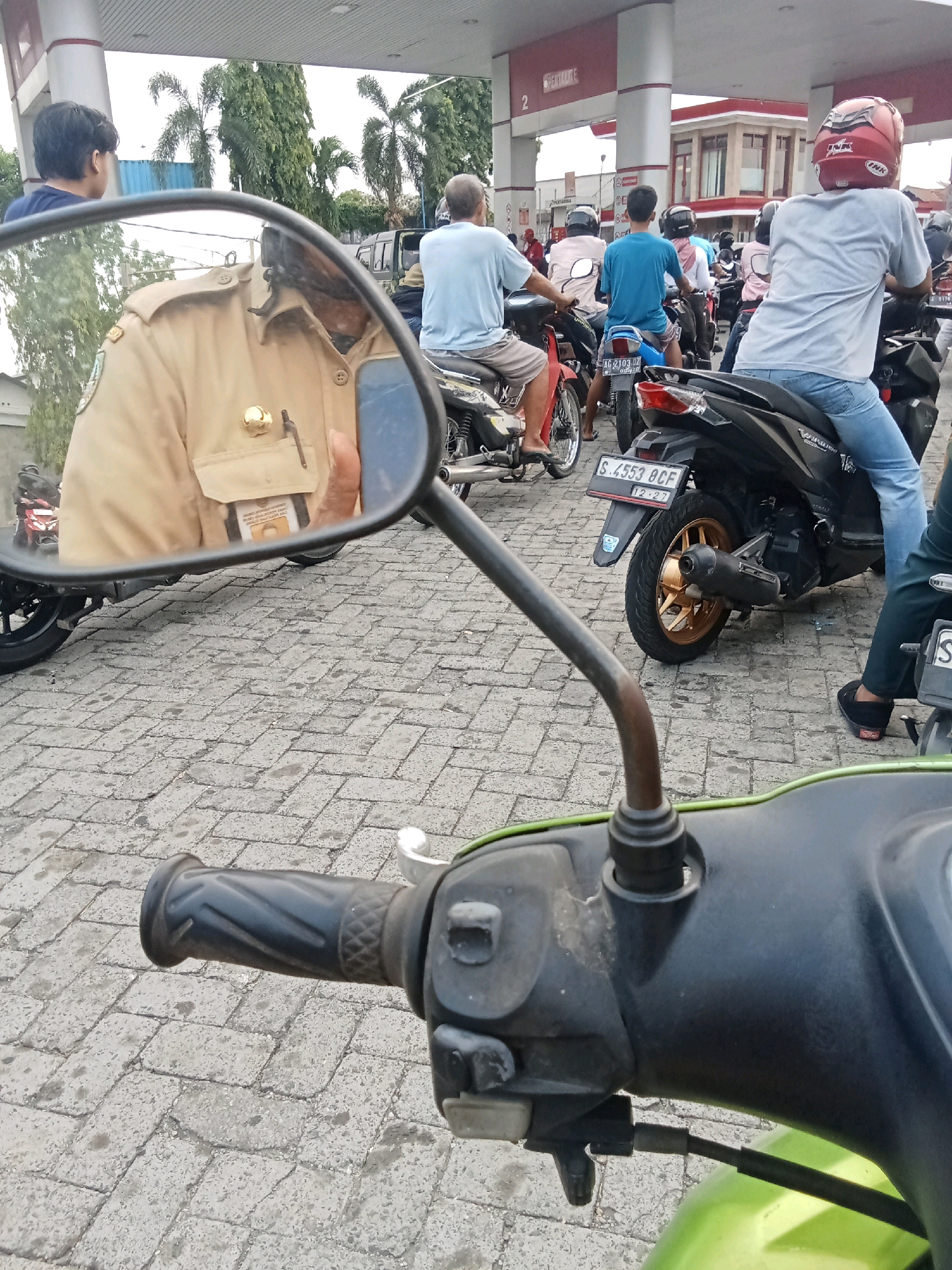 In Jelakombo, Indonesia, a perspective shot from the handlebars of a green motorcycle captures a queue of people on motorcycles at a gas station. The foreground shows the motorcycle's handlebars and part of its body. A rearview mirror reflects a person in a tan uniform, possibly a security guard or government employee, holding a smartphone. Behind the mirror, other motorcycles and riders are visible, some wearing helmets and casual attire. The gas station's white and red structure with a number "2" sign is in the background, along with palm trees and buildings. The ground is paved with grey interlocking bricks. The overall scene depicts a typical day at a gas station in a Indonesian town.