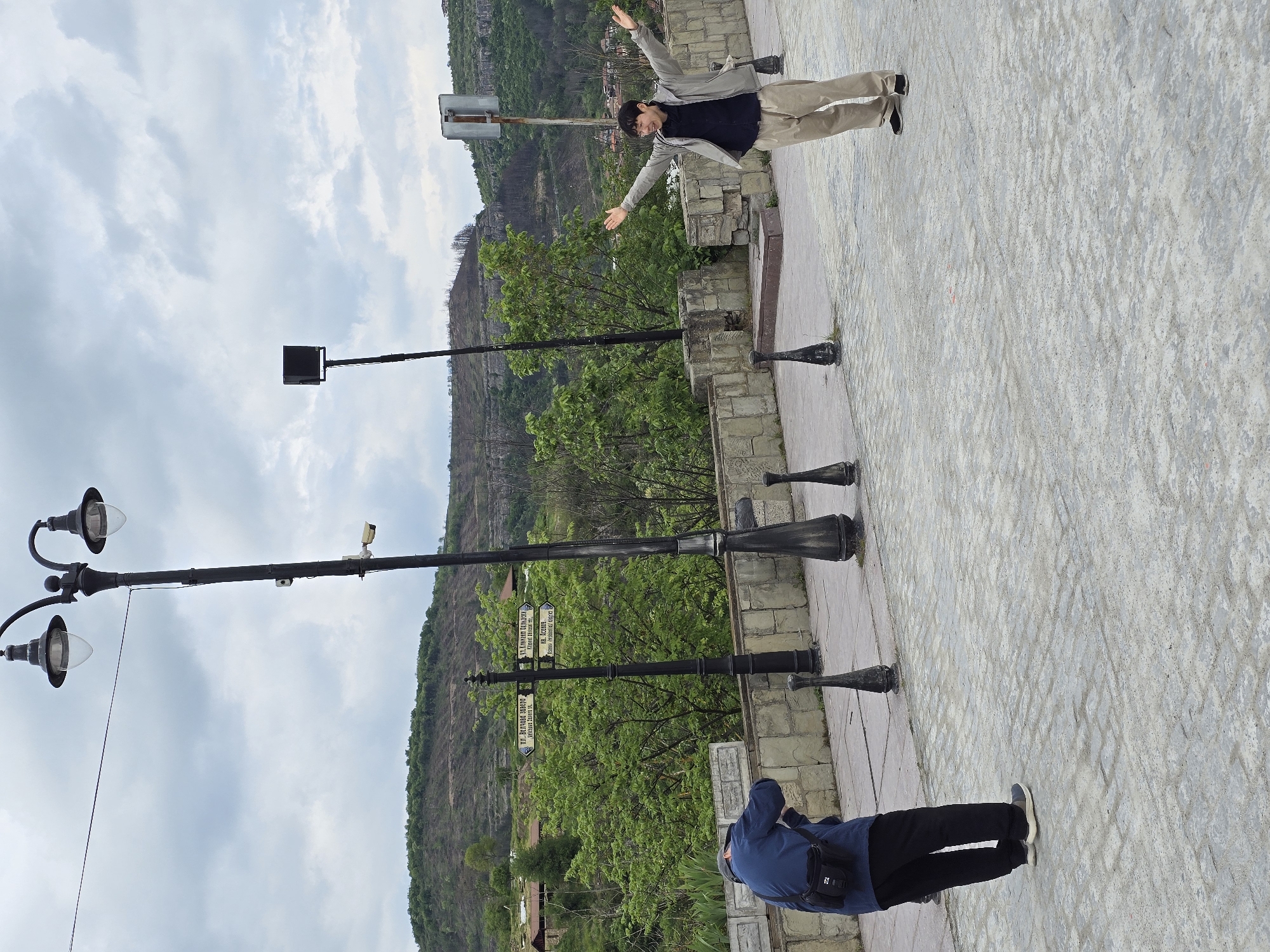 The image shows two individuals on what appears to be a stone walkway or plaza. One person, wearing a light grey jacket, dark shirt, and khaki pants, has their arms outstretched. They are standing near a short stone wall. The other individual, wearing a blue jacket and dark pants, is closer to the bottom of the image and has their head tilted down, possibly looking at something or taking a photo. Several black, vintage-style lampposts are placed along the walkway. Behind the walkway is a green, tree-covered hill or mountain. Street signs are visible on one of the lampposts. The sky is cloudy. The location is unknown.