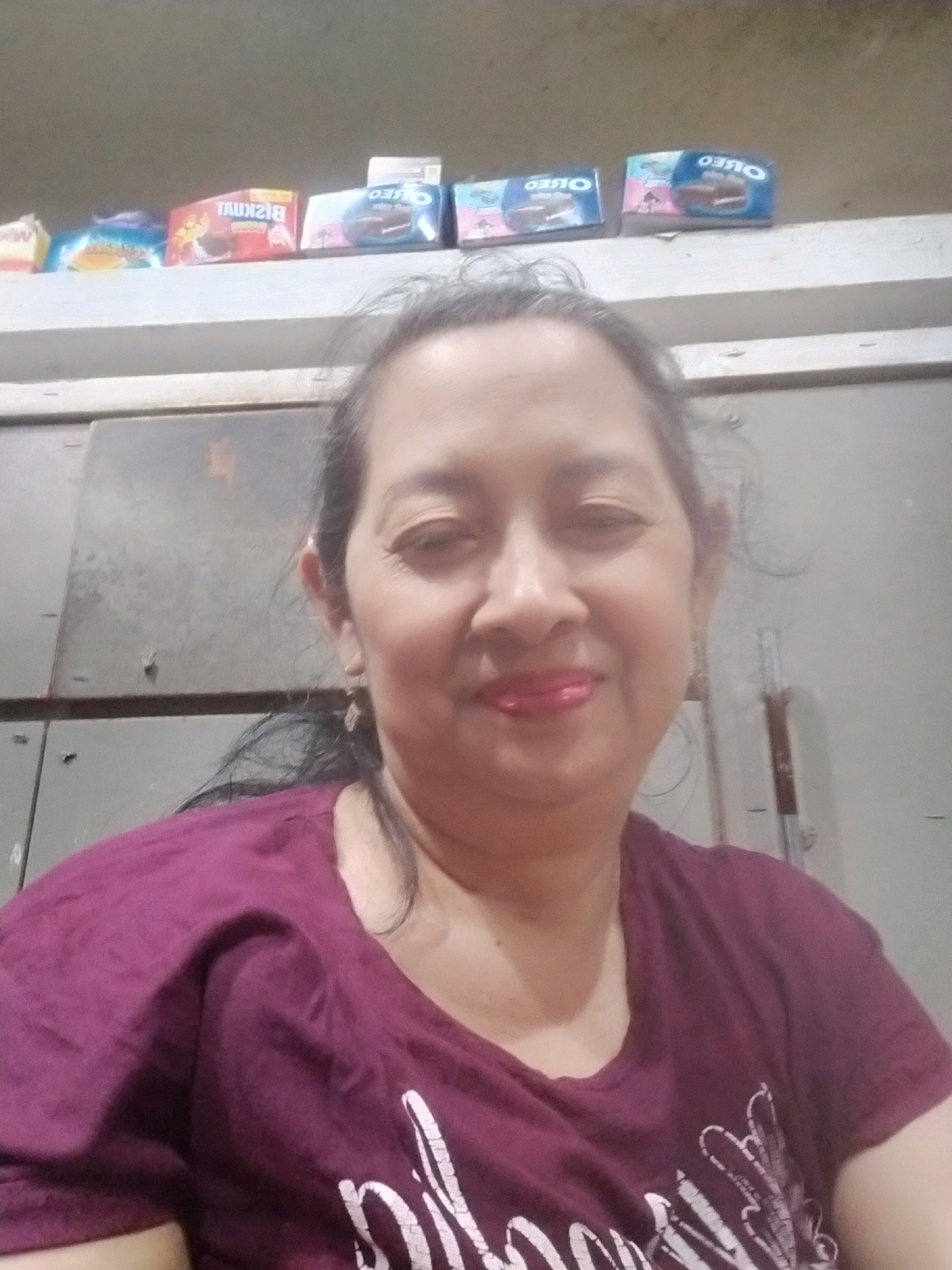A middle-aged woman of Asian descent, with dark hair and wearing a purple t-shirt, poses for a selfie in an indoor setting. The background consists of gray metallic cabinets, a white shelf displaying several snack packages (including Oreo cookies and a package labeled "BISKUIT"), and a neutral-colored wall. The image appears to be taken in Mojodanu, Indonesia.