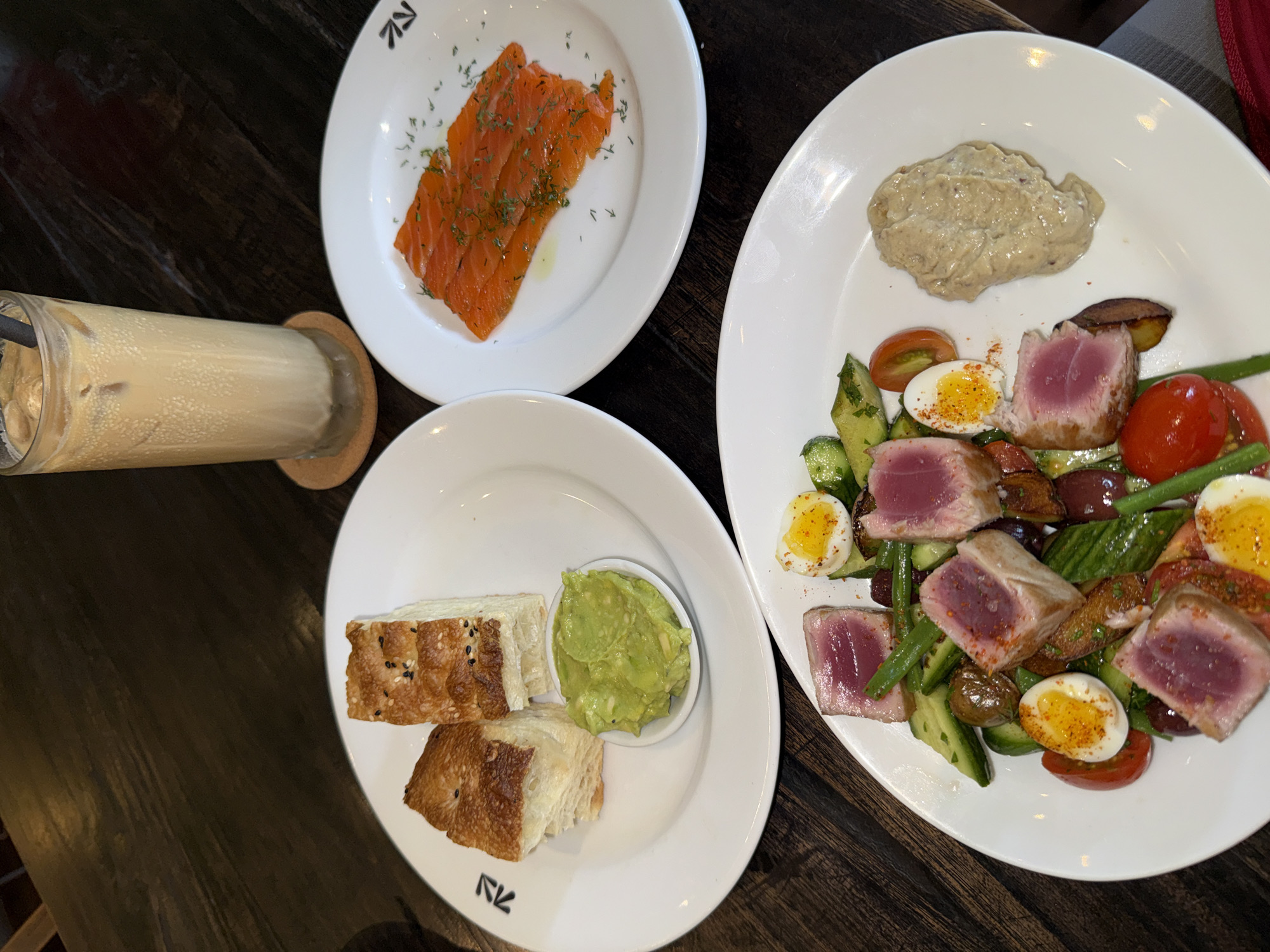 The image shows a table setting with three white plates and a glass of iced coffee. One plate contains smoked salmon garnished with dill. Another plate holds slices of bread with sesame seeds and a small bowl of guacamole. The largest plate features a salad with seared tuna, quartered hard-boiled eggs, green beans, cucumbers, and tomatoes, topped with a scoop of hummus. The table appears to be made of dark wood. The plates have the same logo in the bottom left corner. The setting is likely in North Kuta, Indonesia based on the presence of the logo on the plates which is known to be located in that city.