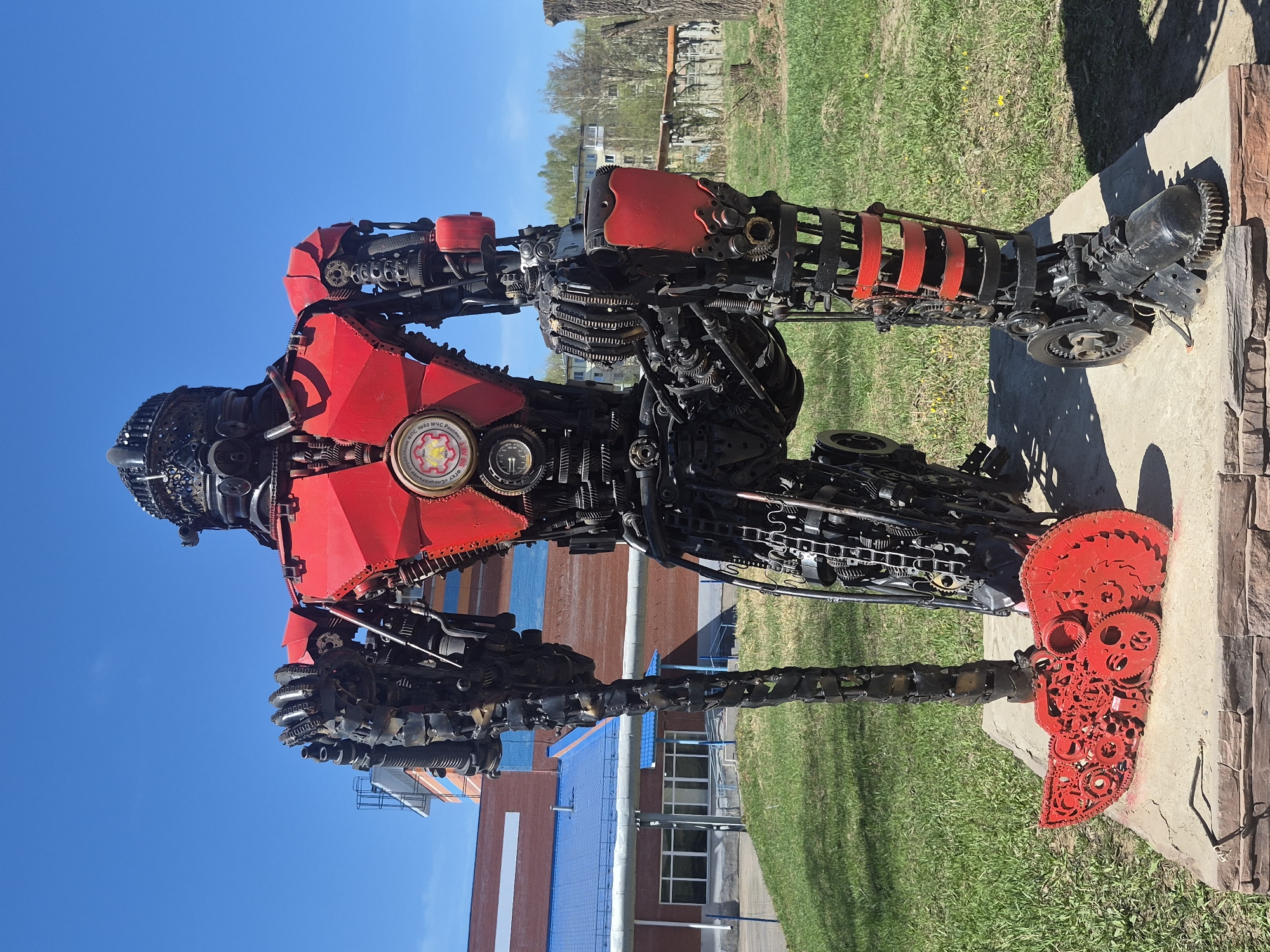 The image shows a sculpture of a robot made from scrap metal and painted red in the city of Votkinsk, Russia. It stands on a concrete plinth in an outdoor setting with grass. The sky is blue and clear. Behind the sculpture are buildings, including one with a red brick facade and blue roof. The sculpture is composed of gears, chains, and other mechanical parts. The "feet" are painted bright red and appear to be formed from large gears. The upper torso of the robot has some red metal plates attached. The background suggests a well-maintained public area.