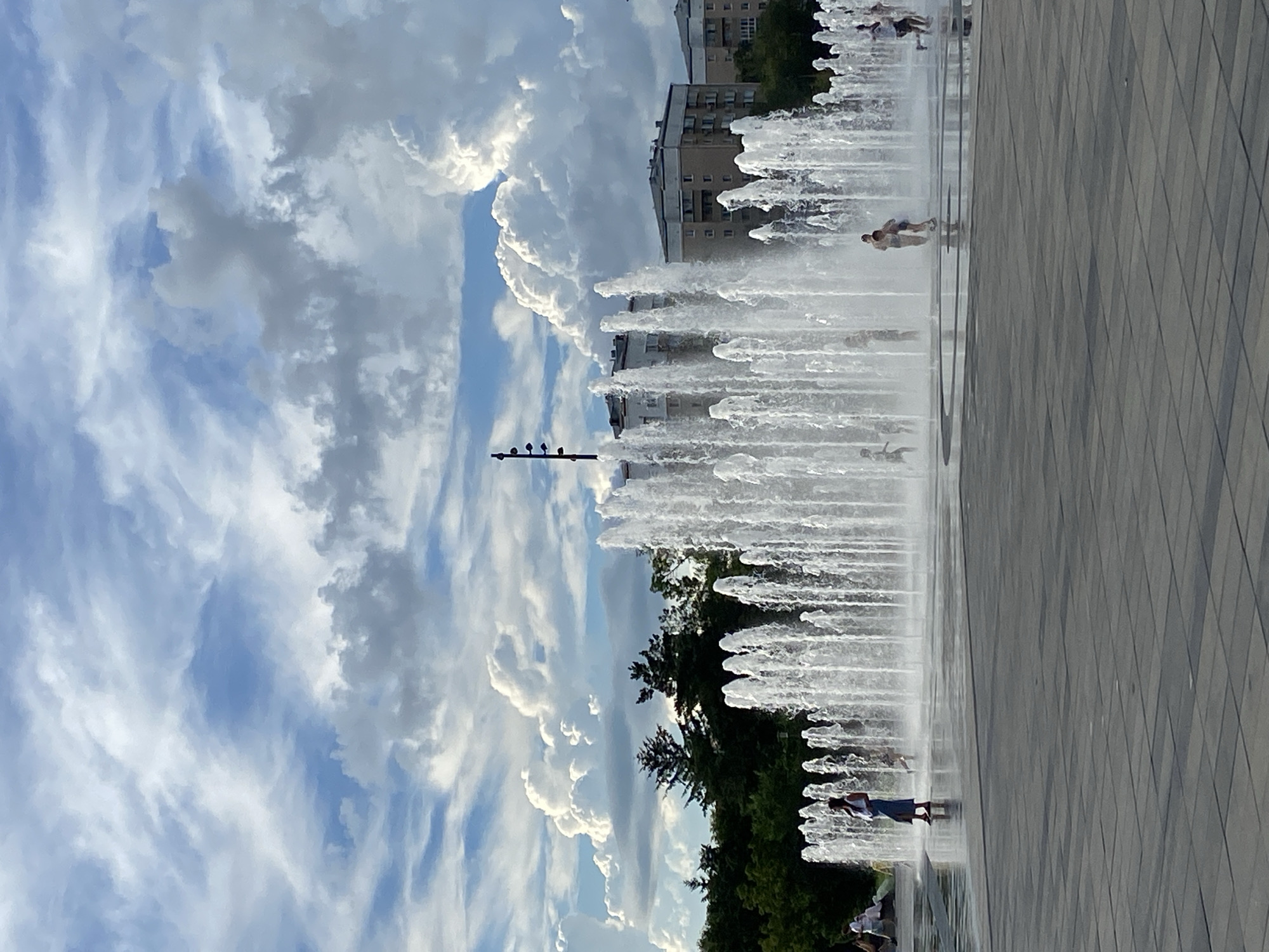 A wide shot captures a vibrant urban scene featuring a large, modern fountain complex with numerous water jets spraying upwards. The fountain is situated on a paved plaza, with several individuals, including children, visible playing in and around the water. In the background, lush green trees frame the scene, behind which stand several multi-story residential buildings. The sky above is a dynamic canvas of blue with dramatic, billowing white and gray clouds, suggesting a lively, possibly summer, day. A single dark lamppost with multiple lights stands tall on the left side of the frame. The overall impression is one of a recreational public space within a city. This is the fountain complex located on Amurskaya Street in Blagoveshchensk, Russia.