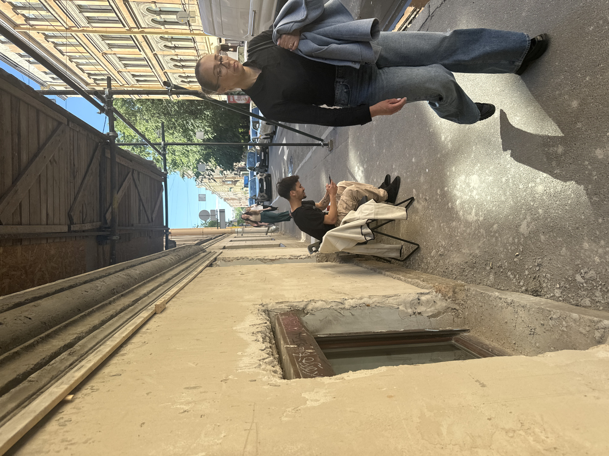 The image shows a street scene in Saint Petersburg, Russia. In the foreground, a woman wearing glasses, a black collared shirt, and wide-leg jeans walks towards the viewer, holding a light blue jacket in her right arm. She is on a sidewalk next to a building with a rough, beige facade and a boarded-up window. Behind her, a man is sitting on a folding chair on the sidewalk, looking at his phone. He is wearing a black t-shirt and light-colored pants. A white cloth is draped over a chair next to him. In the background, there is a street with cars, trees, and buildings with yellow facades. The buildings appear to be in a classical architectural style. A scaffolding structure is visible above the sidewalk, suggesting ongoing construction or renovation. The sky is blue and clear.