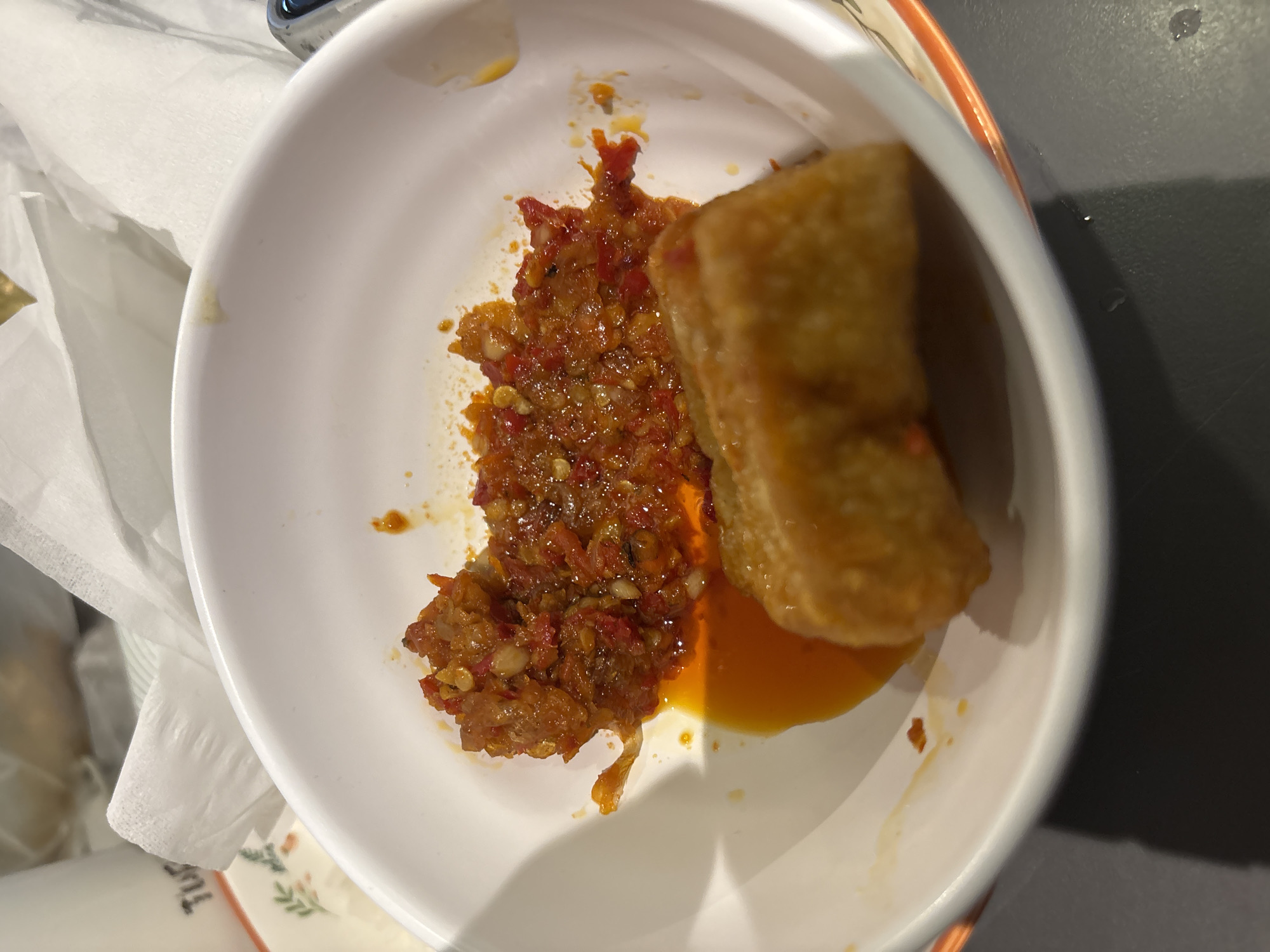 A white ceramic bowl with an orange rim holds a serving of chili sauce and two fried items. The chili sauce is a coarse paste with visible pieces of chili and possibly garlic. A pool of orange oil surrounds the chili paste. In front of the chili paste are two golden-brown fried pieces that appear to be some sort of savory snack or appetizer, possibly spring rolls or battered tofu. The bowl is placed on a dark gray surface, likely a table. In the background, a white paper napkin is visible, and part of a white container with the word "Tur" printed in black is also present. The scene is indoors, and the lighting suggests it is daytime. The overall context suggests a dining setting, likely in a restaurant or home in Pekanbaru, Indonesia, given the details provided.