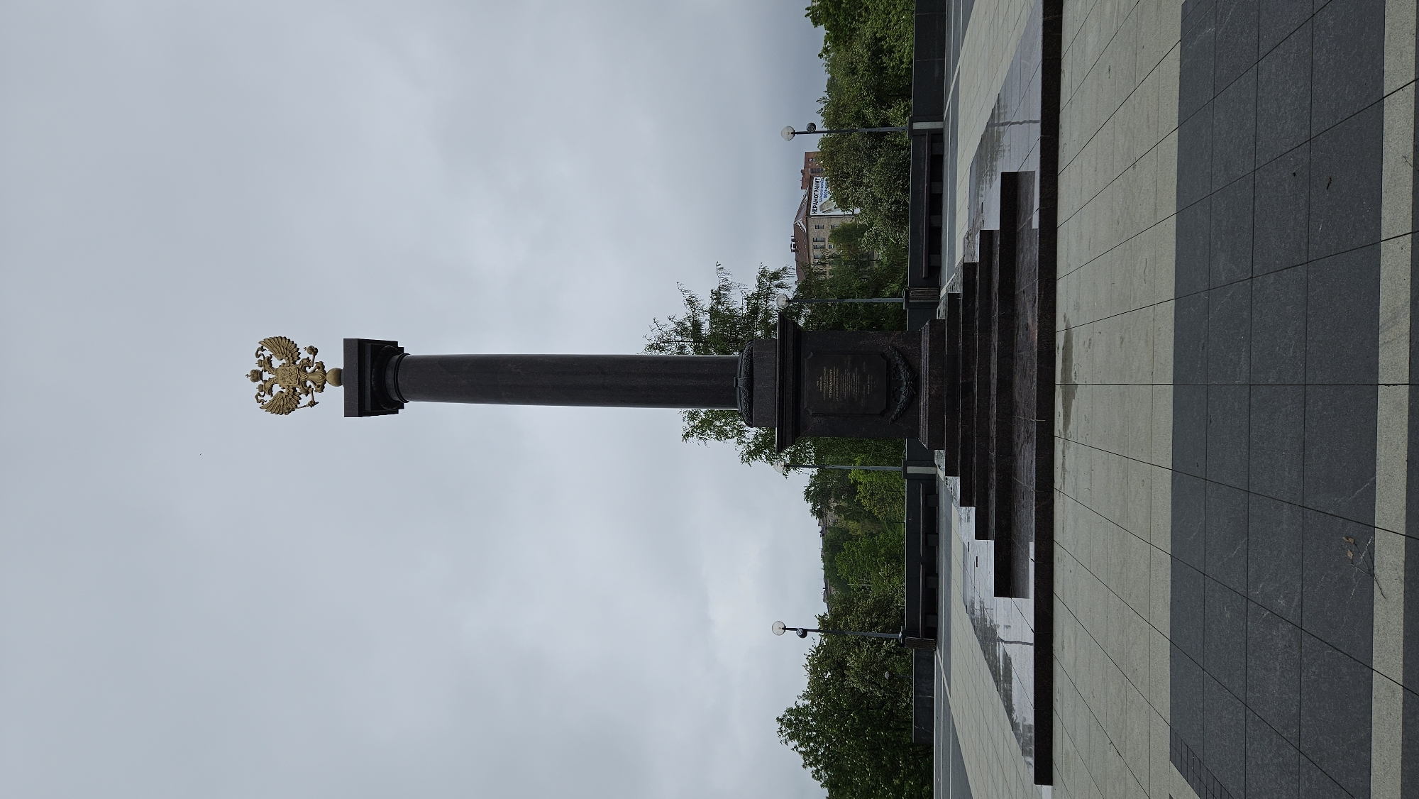 A monument stands against a cloudy sky in an urban setting, city of Unknown, Unknown. The monument, a dark, possibly bronze or stone column topped with a gilded double-headed eagle, is centered in the image. It rests on a multi-tiered base. A plaque is fixed on the base. A paved area, composed of alternating light and dark tiles, extends into the foreground, partly wet. Trees and a distant building with a visible banner are visible in the background. The scene is still and devoid of human activity.