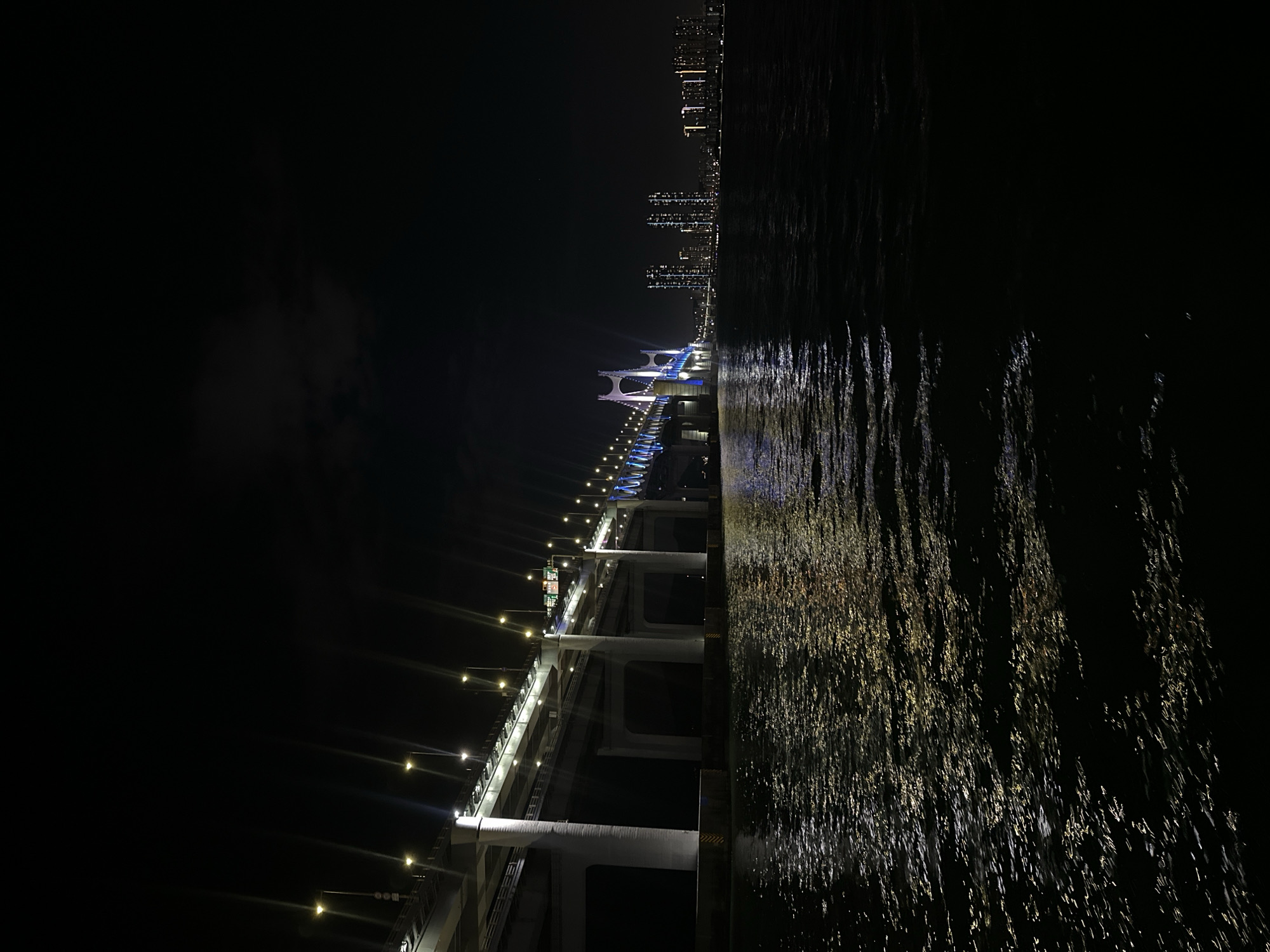 A night-time, low-angle, wide shot captures a brightly lit suspension bridge spanning across a dark body of water. The bridge is illuminated with white and blue lights, creating a striking contrast against the night sky. On the left side of the frame, another elevated road, also lit by streetlights, runs parallel to the water. The water's surface reflects the lights from the bridge and the distant city skyline, creating shimmering patterns. The skyline, visible in the background on the right, features several high-rise buildings with illuminated windows, suggesting a densely populated urban area. The scene is characteristic of the Gwangan Bridge in Busan, South Korea, a famous landmark known for its vibrant night-time illumination. There are no people visible in the image.