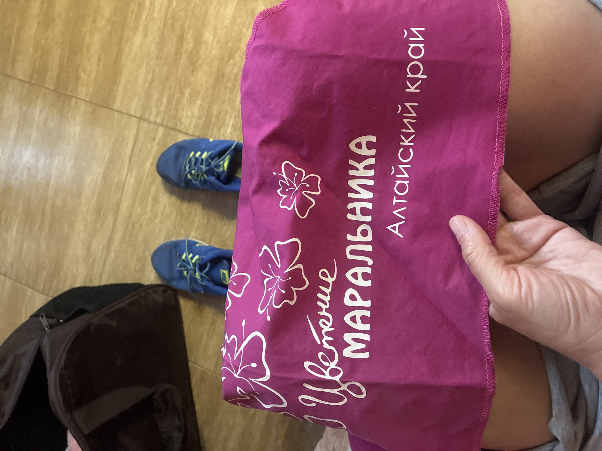 The image shows a person holding a magenta fabric with white lettering and floral designs. The text on the fabric reads "Цветение МАРАЛЬНИКА" and "Алтайский край." In the background, a dark duffel bag is visible next to a pair of blue athletic shoes on a wooden floor. The person's legs are visible from the knees down, with the fabric draped over them. The scene appears to be indoors, possibly in a home or hotel room. There is no indication of the city of Sochi, Russia, in the image.