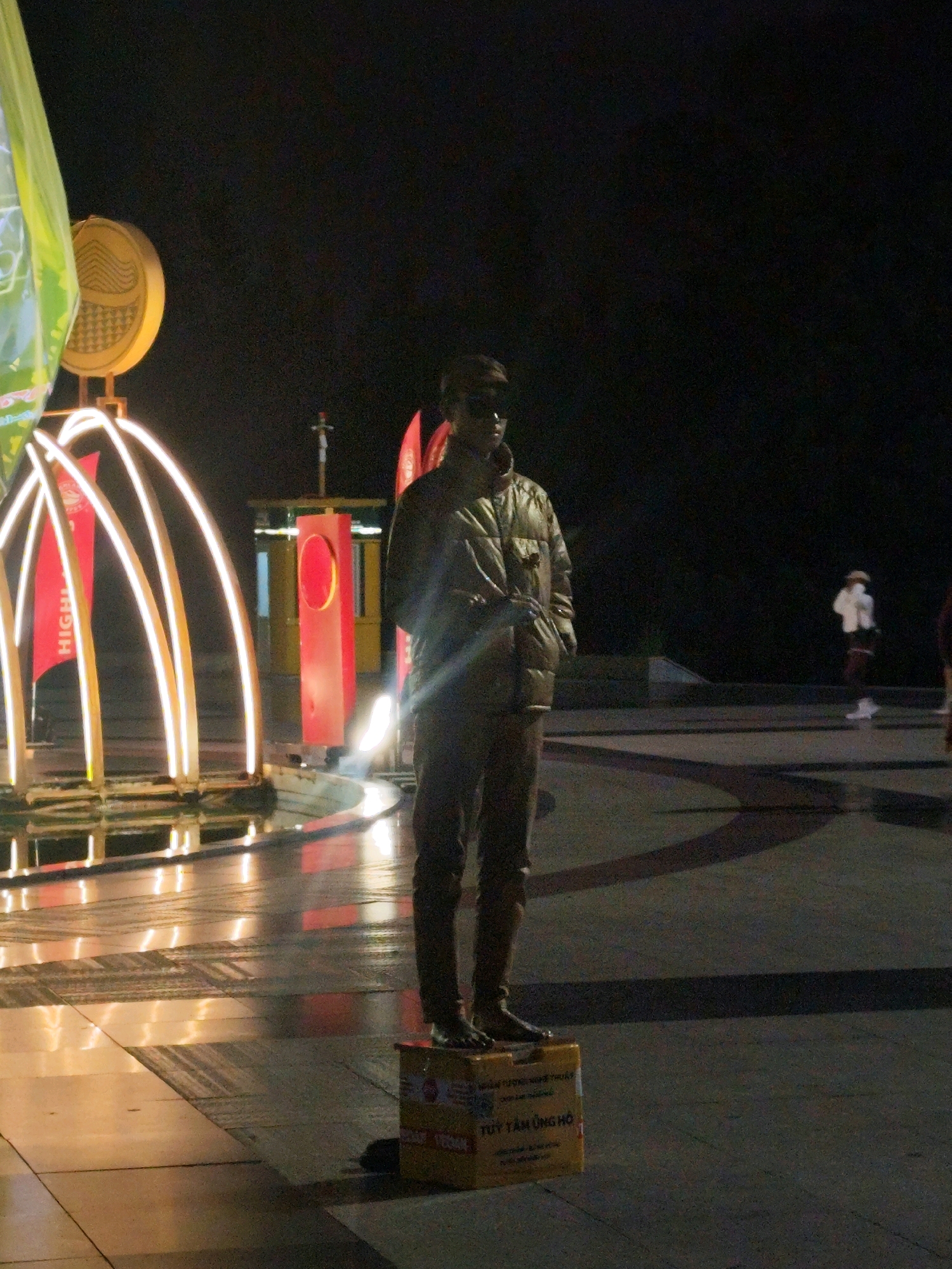 The image depicts a statue of a person standing on a cardboard box in what appears to be a public space at night, possibly a pedestrian area or plaza in Da Lat, Vietnam. The statue is dressed in a puffer jacket and sunglasses, and it is positioned in the foreground. In the background, there are several illuminated structures, including curved lights resembling a fountain or art installation, and some red flags with text, one of which reads "HIGH". Further in the background, other figures, some blurred and distant, are visible, suggesting the presence of other people in the area. The ground is wet and reflects the surrounding lights. The overall atmosphere is dimly lit, characteristic of an evening scene in an urban environment. The cardboard box beneath the statue has text in Vietnamese, which translates to "TUY TAM UNG HO", likely a donation box or for collecting contributions.