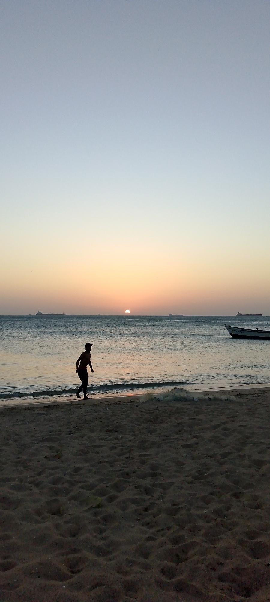 A silhouetted figure runs along a beach at Villa Marina, Venezuela, during sunset. The sun is setting on the horizon, with several ships visible in the distance. A small boat is anchored near the shore. The foreground consists of a sandy beach. The sky transitions from light blue to orange near the horizon.