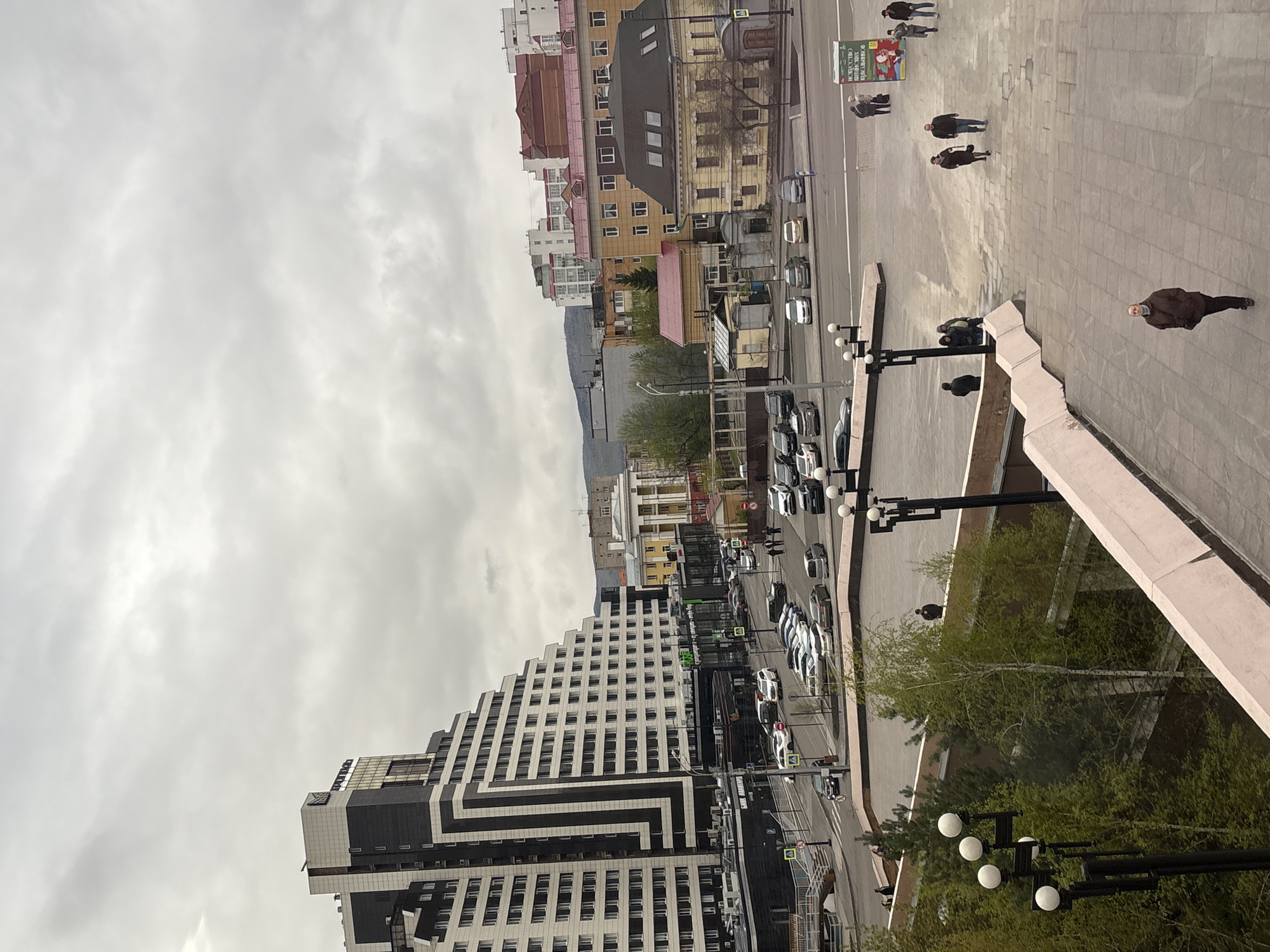 The image shows an urban scene in an unknown city. The perspective is an elevated view looking down onto a street and adjacent pedestrian area. A tall, modern building with a striking geometric design of white and black facades dominates the lower left of the frame. A street runs alongside this building, filled with parked cars and passing traffic. In the center of the frame, further down the street, are older buildings with varied colors and architectural styles. A pedestrian walkway is on the right side of the image, featuring paving stones and a small number of people walking. Trees and street lights border the sidewalk. The sky is overcast and gray, suggesting a cloudy day.