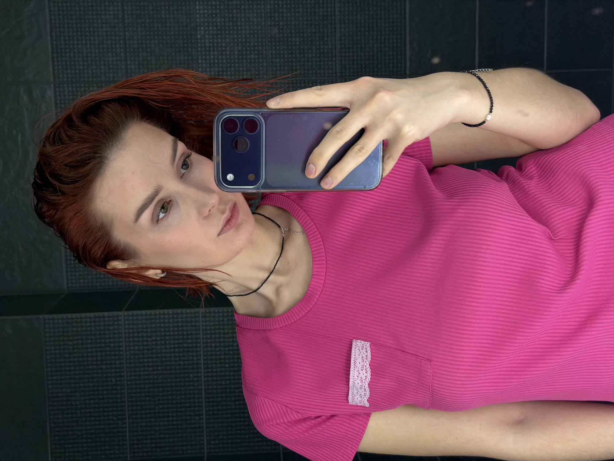A red-haired woman in a pink shirt takes a selfie with a smartphone. The background appears to be a wall with a textured, dark gray tile pattern. The location, Chaikovsky, Russia, is not discernible from the image alone.