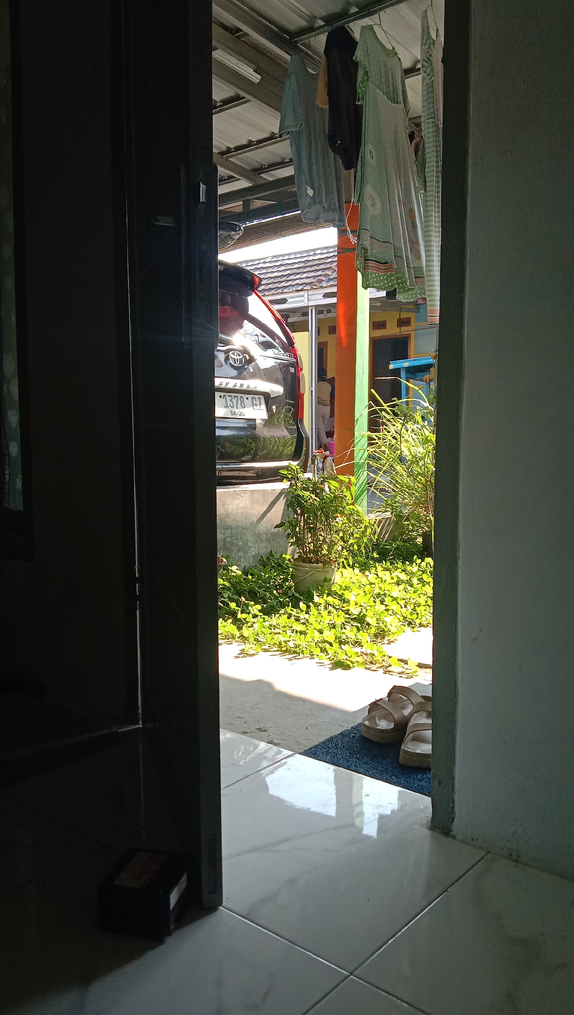 The image shows an outdoor scene viewed from an interior doorway. In the foreground, a dark doorway on the left and a white wall on the right frame the view. The ground is tiled with large, light-colored squares reflecting sunlight. To the right, a blue mat holds a pair of beige sandals.
Beyond the doorway, a black car, identified by its license plate as "B 1378 GT" and a "04-26" sticker, is parked. The car has a Toyota logo on its rear. In front of the car, there is a concrete planter box filled with lush green ground cover plants. A taller potted plant with green leaves is situated behind the ground cover. Sunlight casts shadows across the scene.
Above the car and plants, a metal roof structure is visible, supported by poles. Laundry hangs on a line beneath this roof, including a light blue t-shirt, a darker t-shirt or shirt, and a light green dress with a floral pattern. A bright orange pole stands to the right of the car. In the background, a building with yellow and orange walls is partially visible, along with a tiled roof. The overall impression is of a residential outdoor space in Karangmangu, Indonesia, likely a carport or a backyard area.