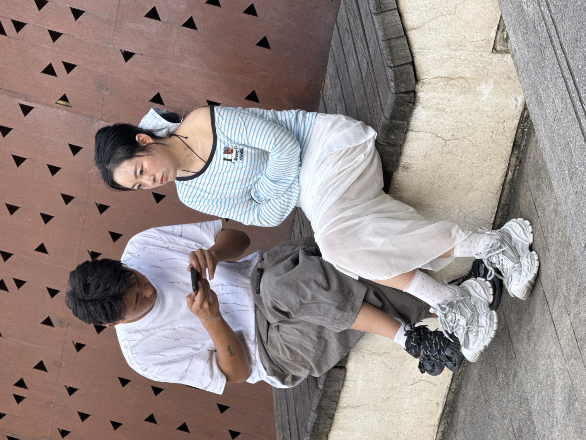 A medium shot depicts two individuals in Yuzhong District, China. One person, likely male, is seated, holding a smartphone in portrait orientation, appearing to take a picture or record video. He wears a white T-shirt with a speckled pattern and gray shorts. Tattoos are visible on his arm. The other person, likely female, is sitting nearby and to the right, wearing a light blue and white striped long-sleeved shirt, a white skirt, and white socks with a subtle pattern. The setting includes a patterned wall with triangular cutouts, wooden planks, and concrete pavement. Their footwear consists of bulky sneakers, with both wearing different styles and colors. The pair are possibly on a park bench or ledge.