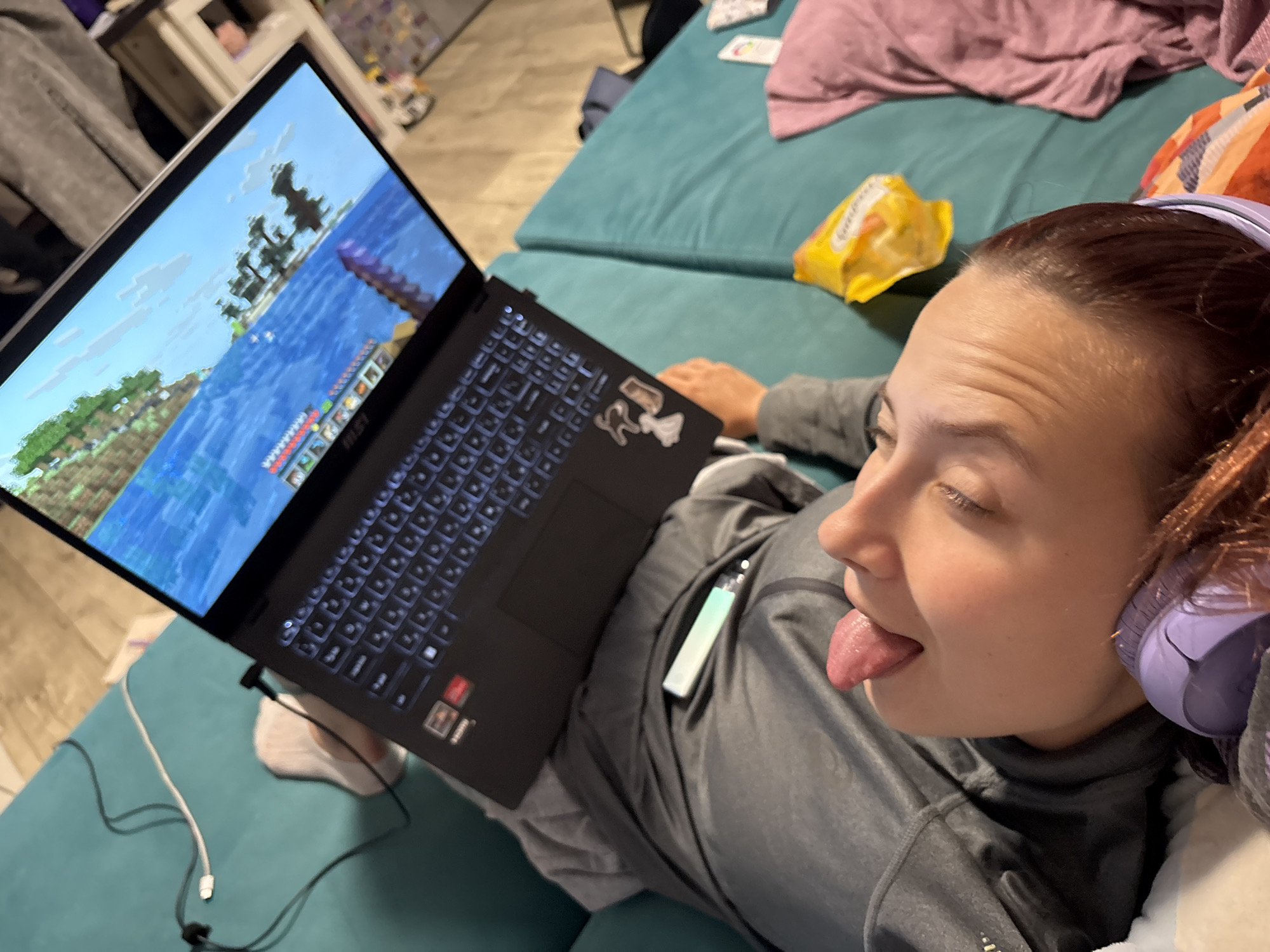 An indoor, high-angle photograph depicts a young adult female lying horizontally on a teal upholstered surface, which appears to be a sofa or bed. Her head is positioned to the right of the frame, angled upwards towards the laptop screen resting on her lap/abdomen. She has medium-dark brown hair, styled back, and wears light purple over-ear headphones. Her eyes are closed or heavily lidded, and her mouth is open with her tongue extended outward. She is dressed in a dark grey long-sleeved top.
A black MSI laptop is open before her, displaying a brightly illuminated screen rendering a scene from the video game Minecraft, featuring blocky green land, trees, and blue water with in-game boats and a visible heads-up display at the bottom. The laptop's keyboard is backlit with blue light, and several white stickers are affixed to its palm rest. A slender vape pen with a light blue-green gradient is visible resting on her chest, just above the laptop.
To the right of her head, on the teal surface, is a crumpled yellow snack bag. Various cables, including white and black ones, are on the teal surface near the laptop. The background is blurred, revealing light wooden flooring, a white cabinet or shelf containing miscellaneous items, and other scattered textiles, including pink and striped fabrics. The lower left of the frame shows a portion of a white sock on her foot.