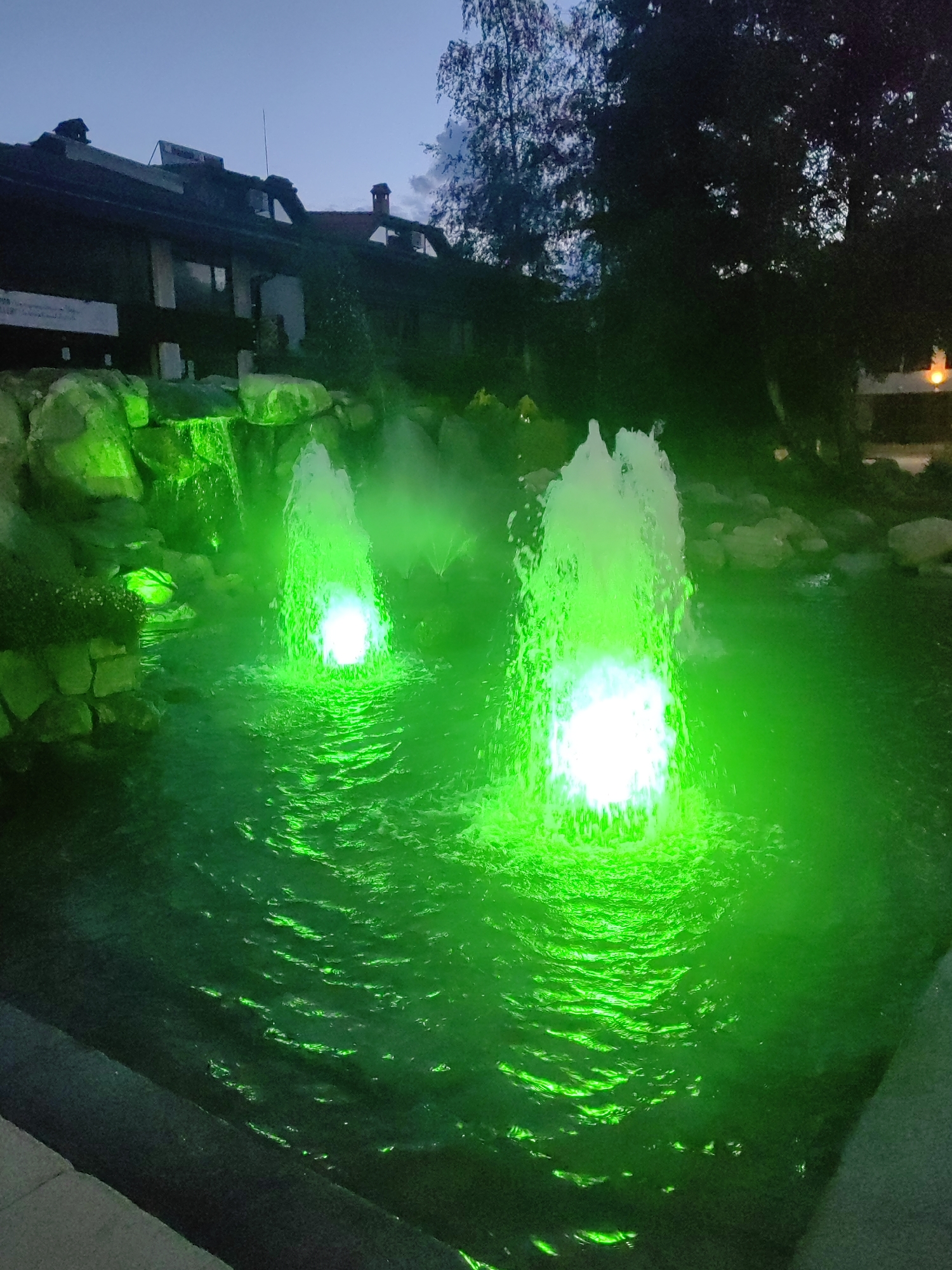 This outdoor scene captures a vibrant water feature at twilight or night. Two prominent fountains vigorously spray water upwards, illuminated from below by intense green lights. The core of the water plumes appears bright white due to the light's intensity, while the surrounding spray and the water in the basin glow a vivid green, creating dynamic patterns and reflections. To the left, a smaller waterfall cascades over rocks, also bathed in the same green light.
The water feature is surrounded by dark, rugged rocks. In the background, a large, dark building with a pitched roof, several windows, and at least one chimney is visible, silhouetted against a dusky blue sky. Some faint, unreadable text appears on banners or signs attached to the building. Dark trees frame the upper right portion of the image, their foliage visible against the sky. The foreground shows a dark, flat edge, possibly a pathway or border for the water installation. The overall impression is one of a decorative, illuminated landscape feature, possibly at a resort or park, designed to be appreciated in the evening. The green lighting evokes a magical or tranquil ambiance.