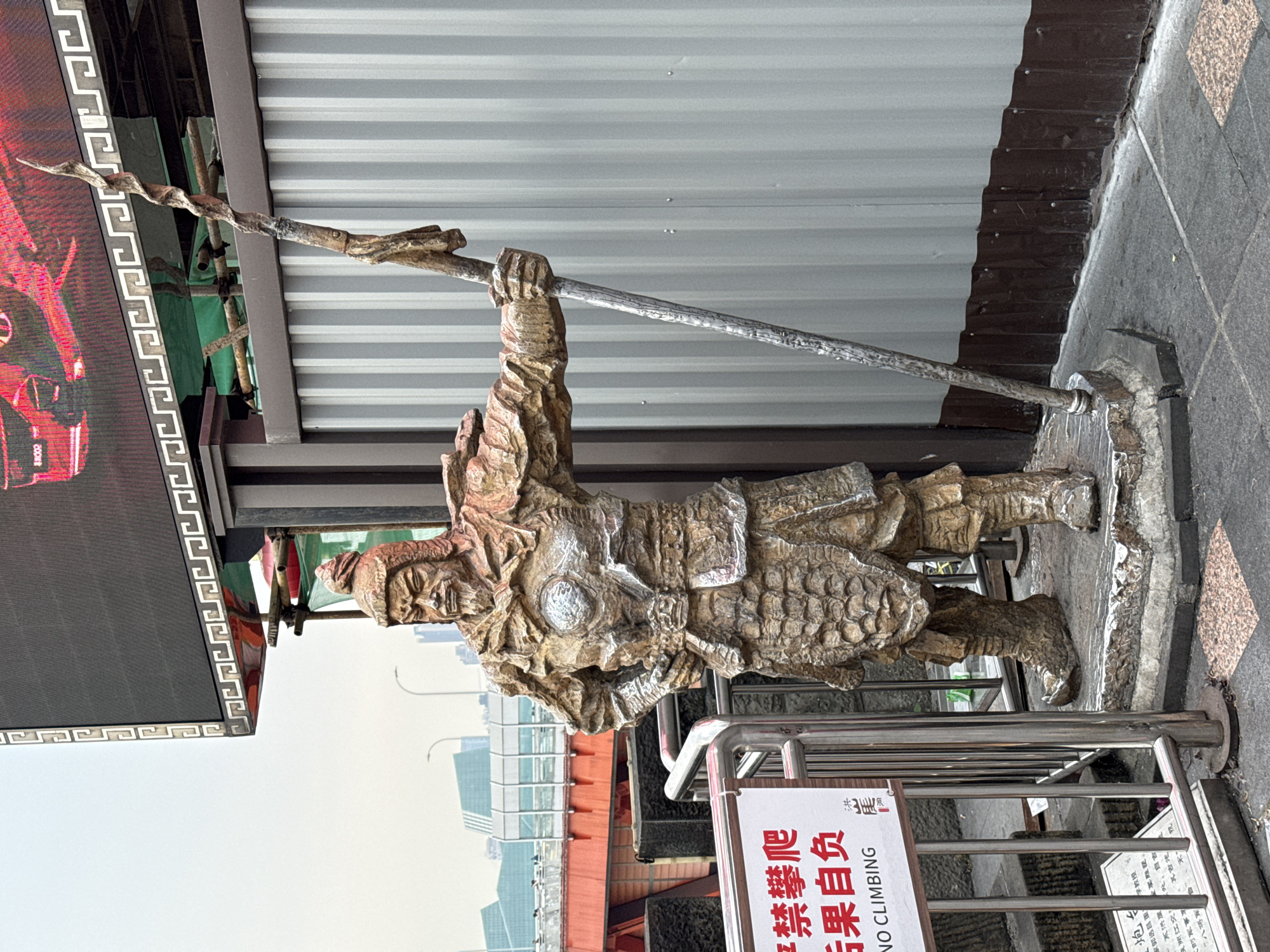 The image depicts a statue of a warrior figure in what appears to be armor, wielding a polearm. The statue is positioned on a raised platform, with a metal railing around it, and is near a sign with Chinese characters and the English phrase "NO CLIMBING". The background includes modern buildings and a grey corrugated metal wall. The statue is in the Yuzhong District of China.