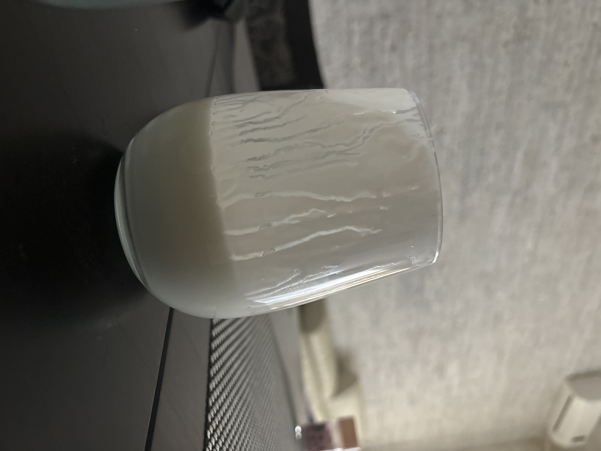 A clear glass with a white, opaque liquid inside rests on a dark surface. The liquid shows streaks or layering patterns within. The surface appears to be next to a woven or textured object. The background includes a white, slightly mottled wall. The precise location within the city of Budva, Montenegro, cannot be determined from the visual data. No people are visible. No specific actions or interactions can be deduced.