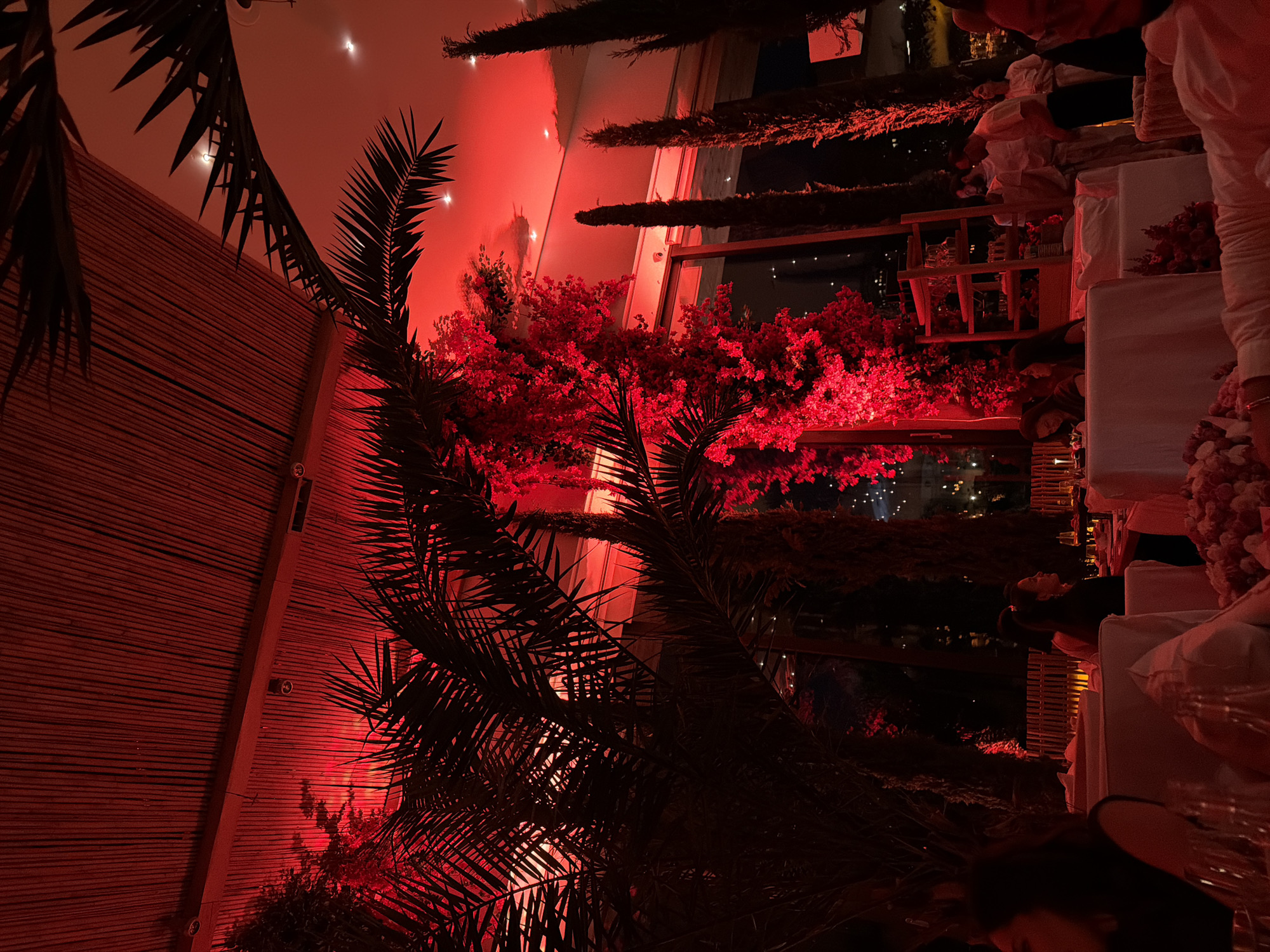 The image depicts a dimly lit indoor dining establishment with a strong red lighting scheme. The ceiling appears to be made of bamboo slats. In the foreground, there are tables set for dining, covered with white tablecloths and adorned with floral arrangements. Palm fronds and other lush foliage, illuminated by the red lights, create a tropical atmosphere. Several people are visible at the tables and standing around, engaged in conversation and dining. The scene suggests a sophisticated social gathering or restaurant setting. The location is identified as being in the city of Yerevan, Armenia.