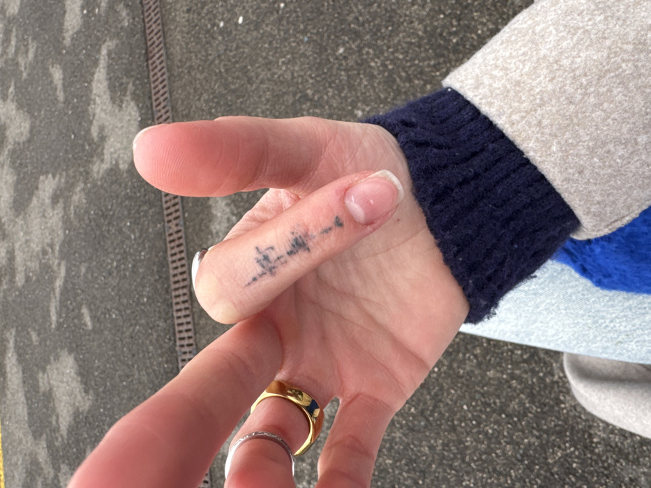 A close-up of a person's hand is presented, displaying two rings, one silver and one gold, on the ring finger. A small black tattoo is visible on the side of the index finger. The person wears a blue sweater with a beige coat. The background shows a section of pavement with a drainage grate.