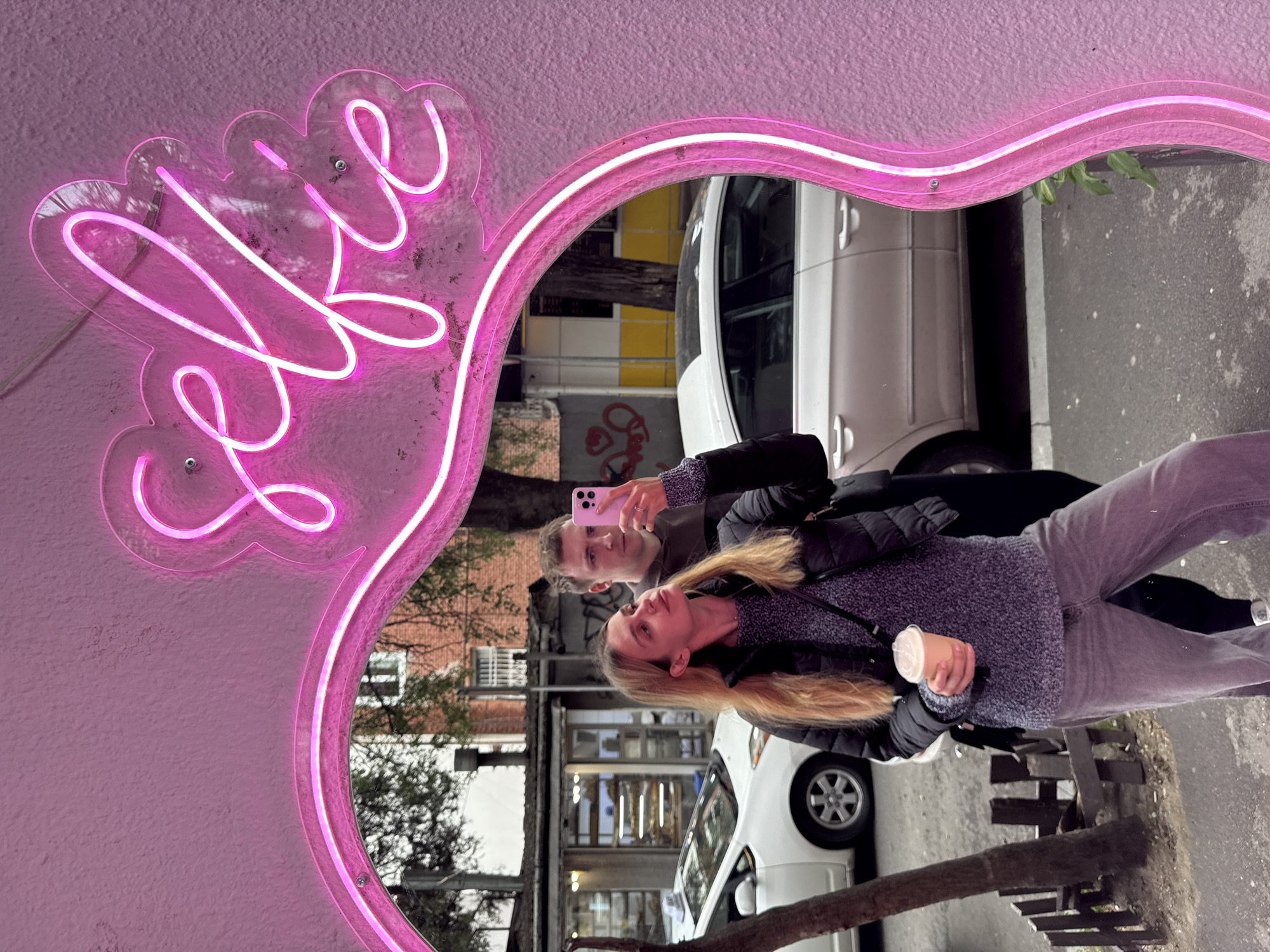 A couple is taking a selfie in the reflection of a mirror with a pink neon "selfie" sign attached, located in Tbilisi, Georgia. The man is holding a pink smartphone. The woman is holding a cup. A white car is parked behind them. The background includes buildings and trees.
